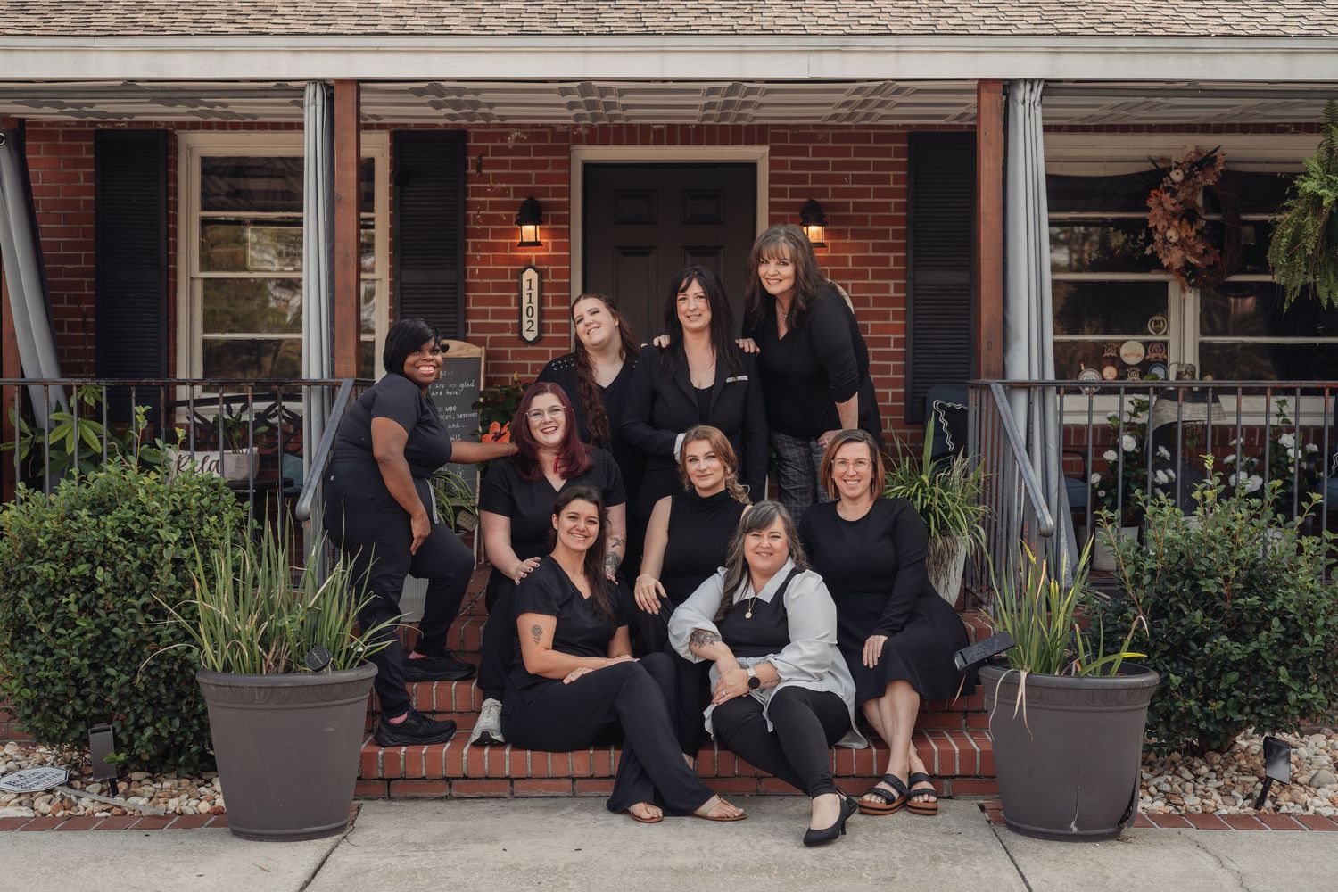 A group of women in black outfits posing together on the front steps of a brick building with potted plants and a dark front door.