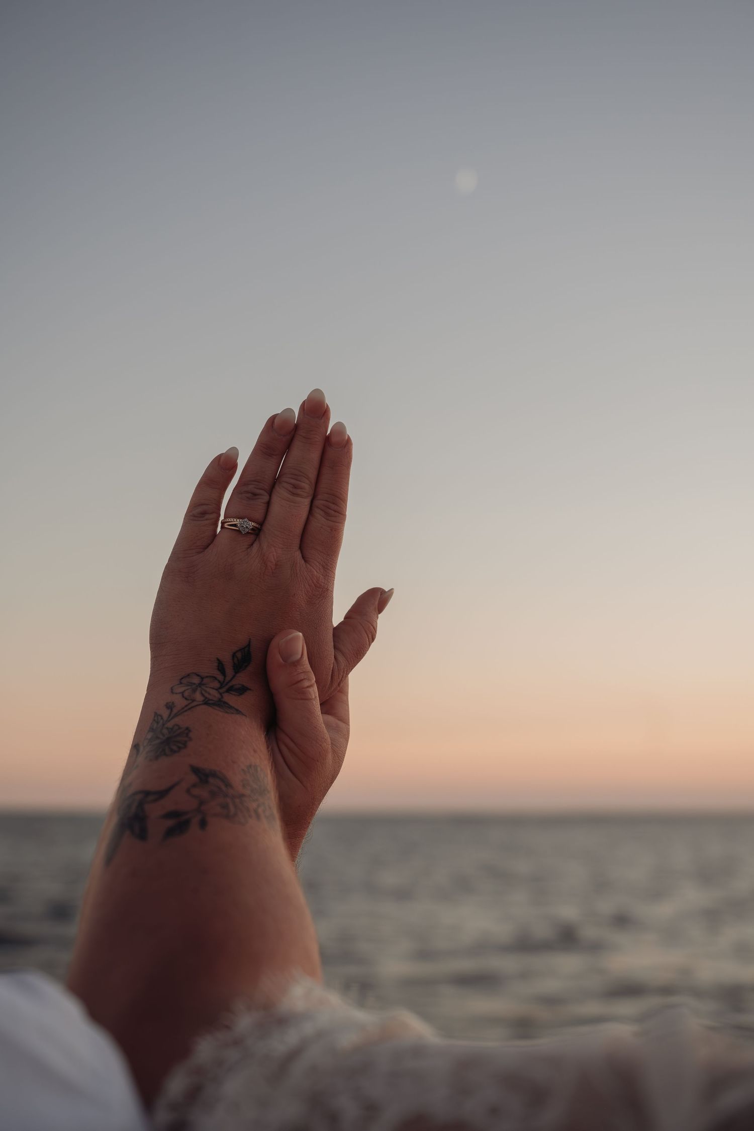 Two hands with a floral forearm tattoo and a ring raised toward a pastel sunset sky above a calm ocean with a faint moon.