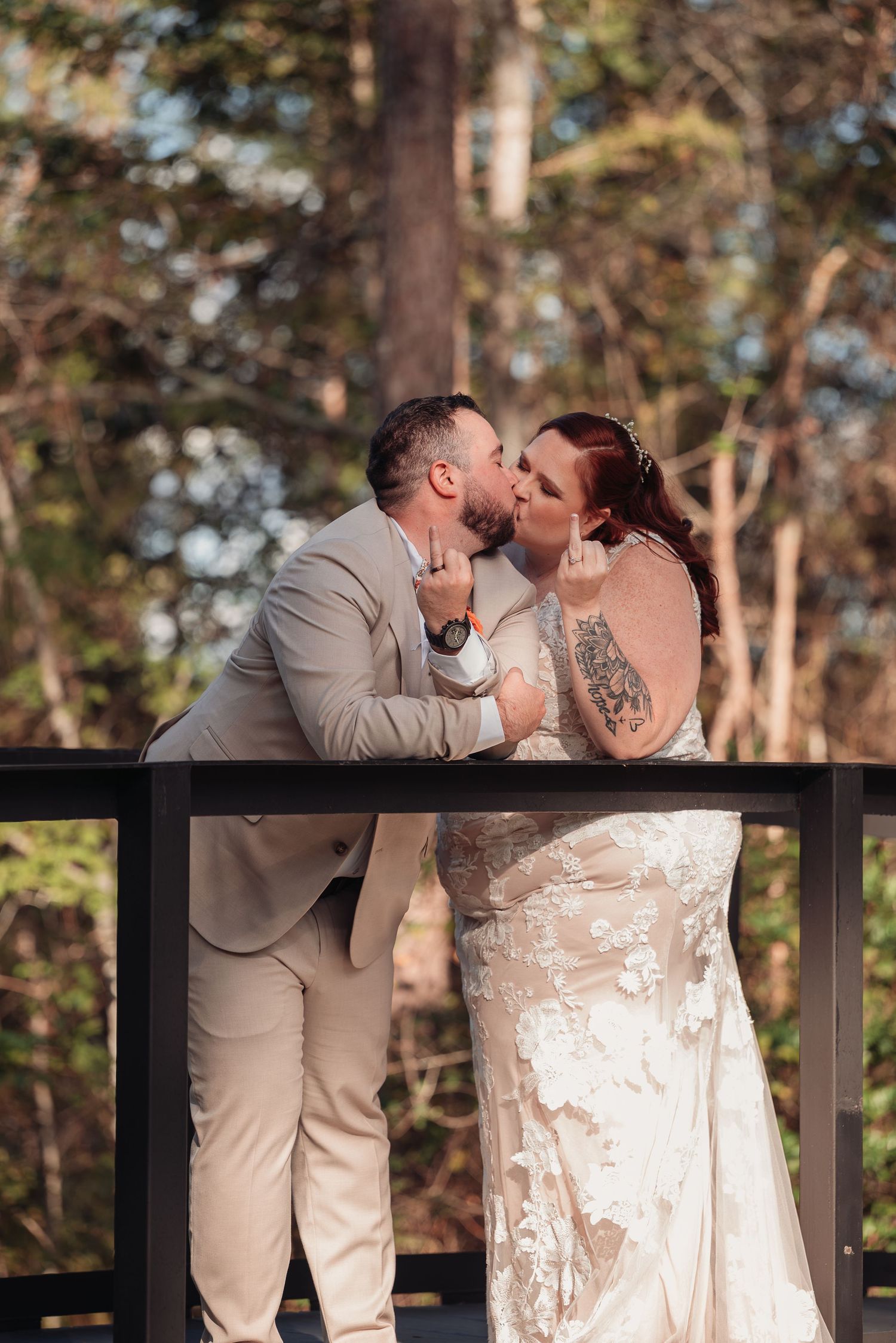 Bride in a lace wedding gown and groom in a tan suit share a kiss on a wooden bridge surrounded by sunlit trees.