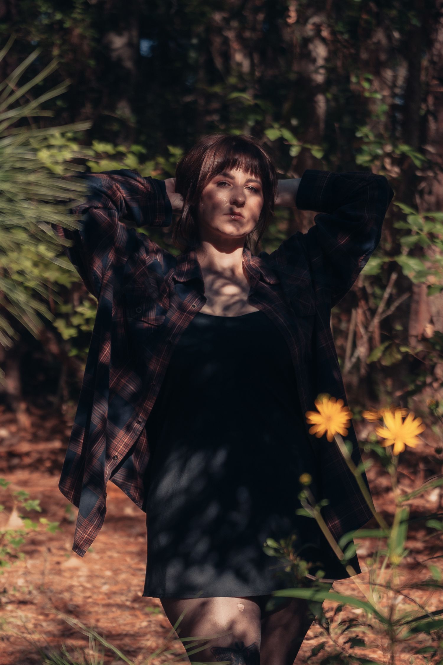 Young woman in plaid shirt and black dress poses in sunlit forest with yellow wildflowers.