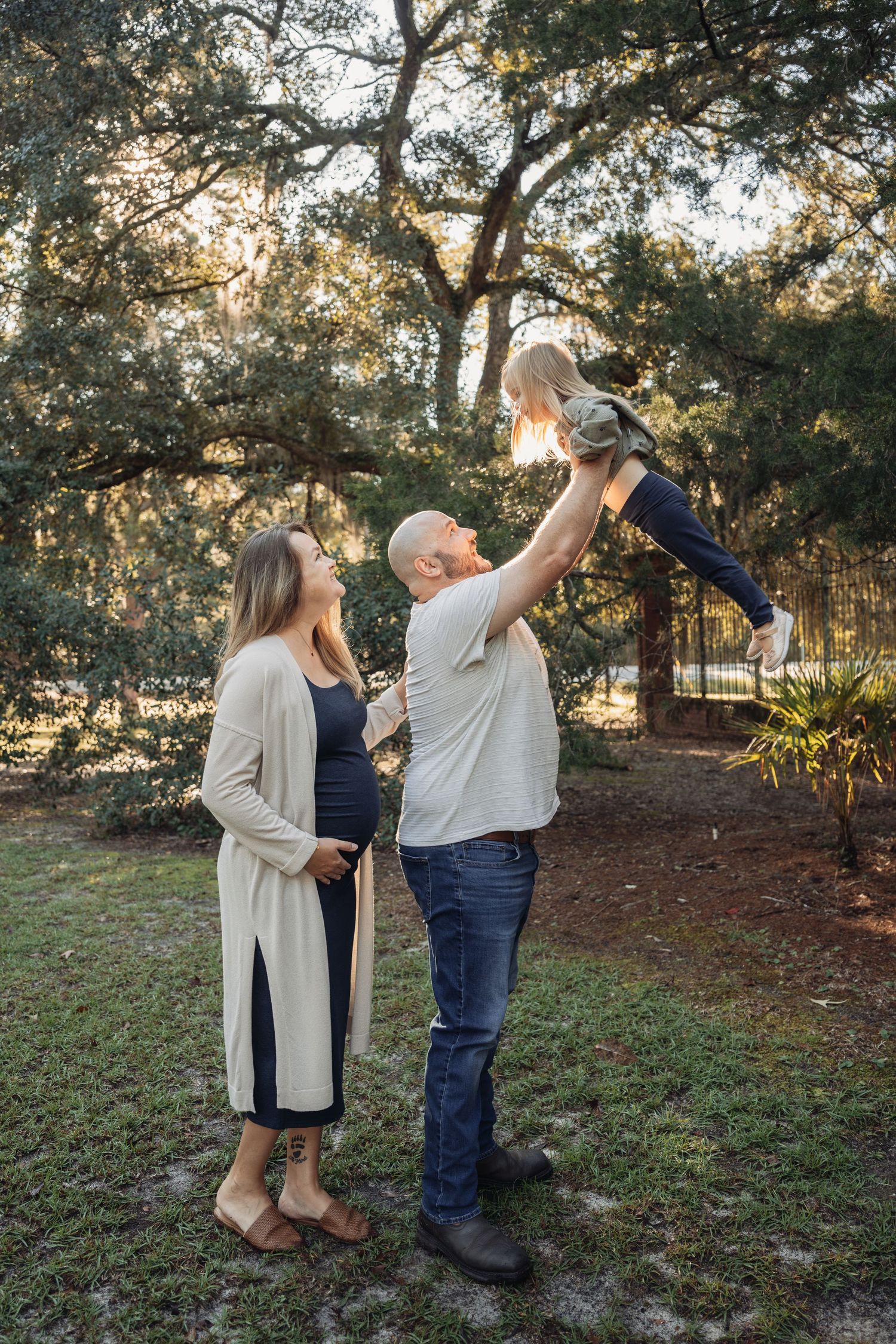Pregnant woman and bald man lifting toddler in sunny park with large oak trees in background.