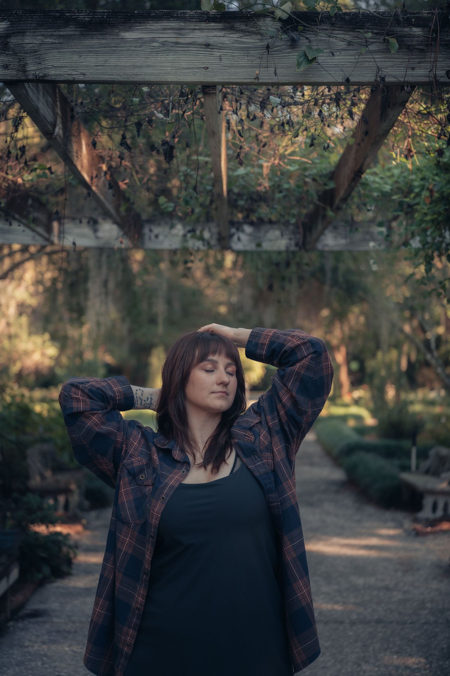 Woman in plaid shirt poses with hands behind head under a rustic wooden pergola in a lush garden setting.