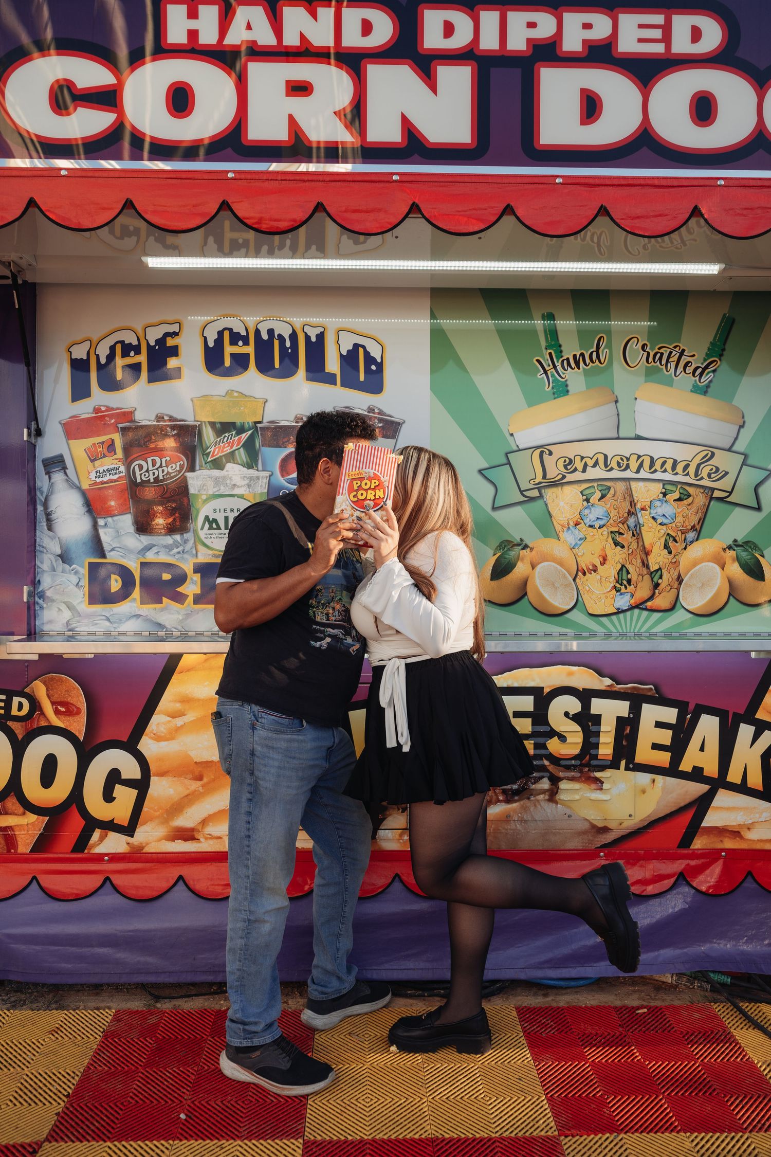 Couple kissing behind a snack held between them in front of a colorful hand dipped corn dog and lemonade fair food stand.