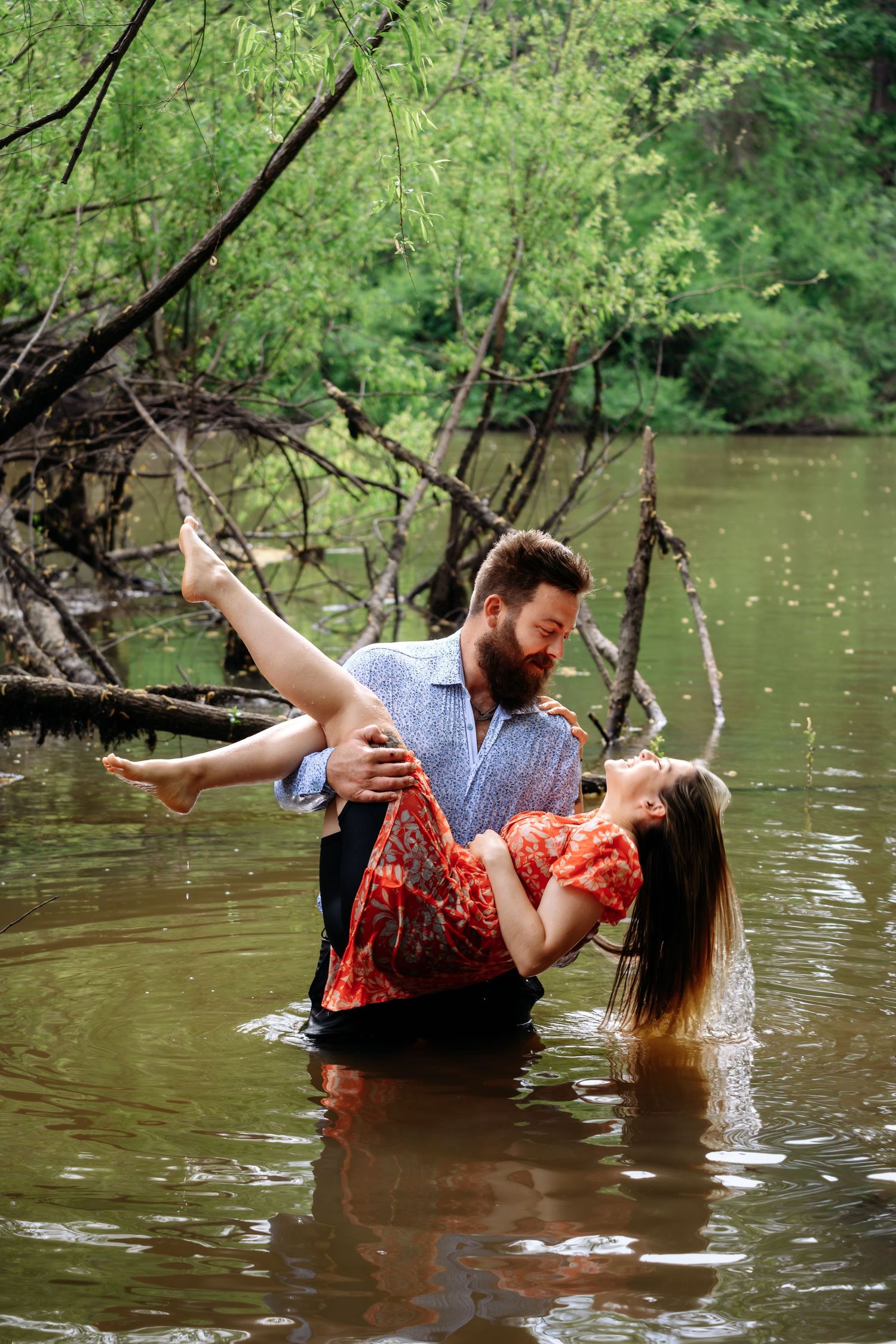 Bearded man carrying his fiancee in his arms while standing in a river outdoors.