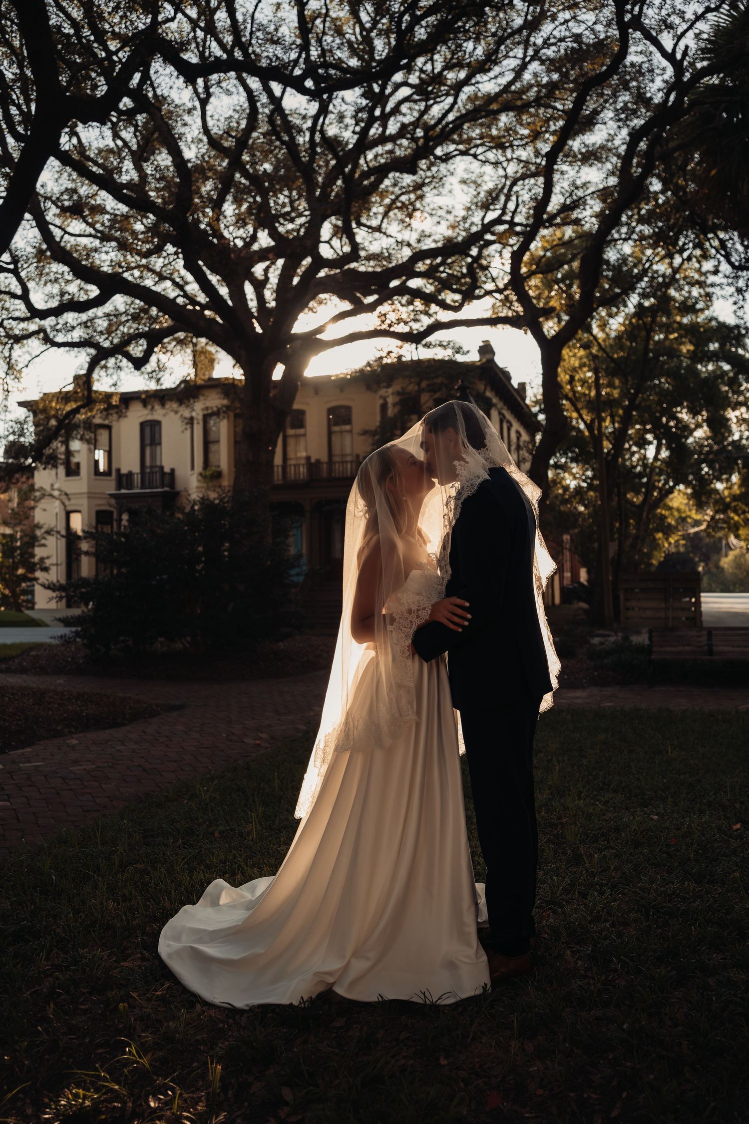 Bride and groom share a romantic kiss under a sprawling oak tree at golden hour in a park.