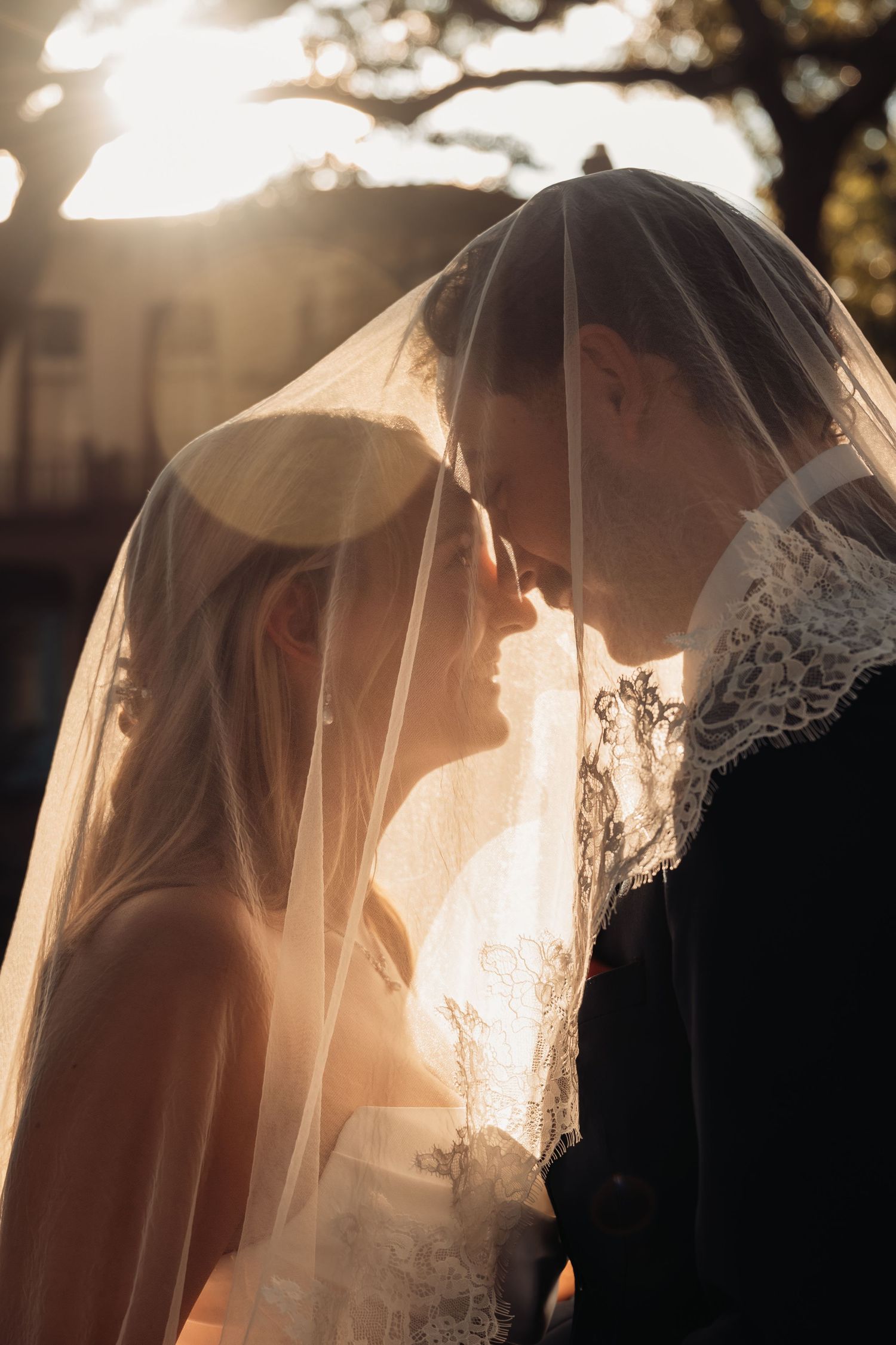 A bride and groom touch foreheads under a lace veil with golden sunlight glowing softly behind them.