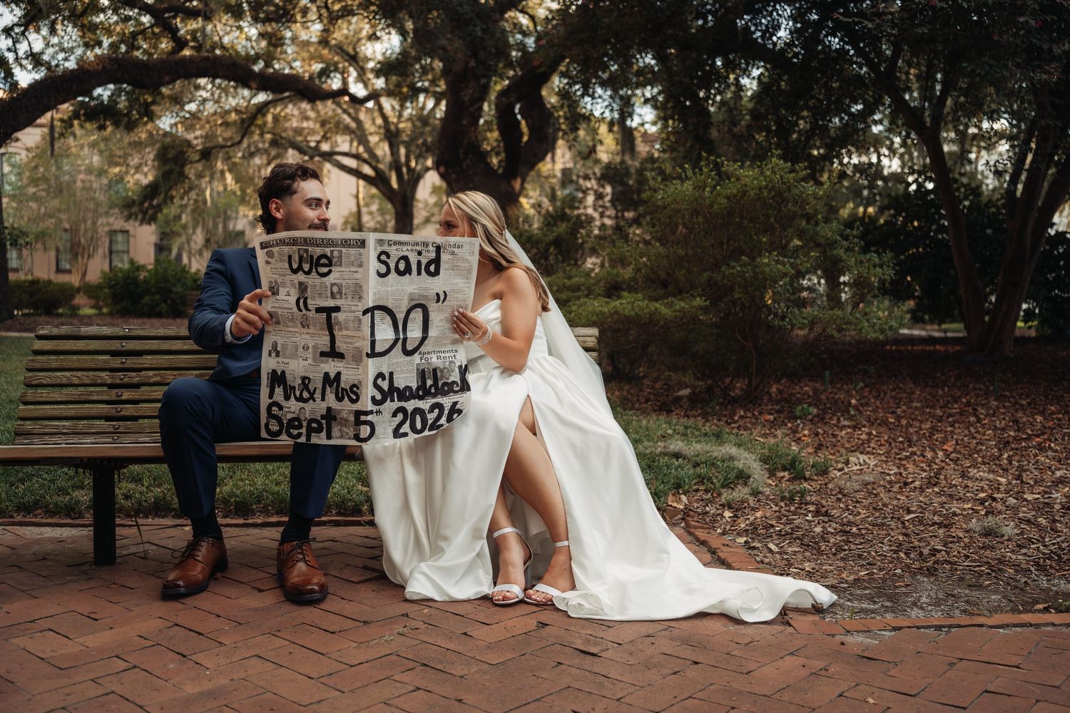Newlyweds sit on a bench holding a sign reading their wedding date, Sept 5 2025.