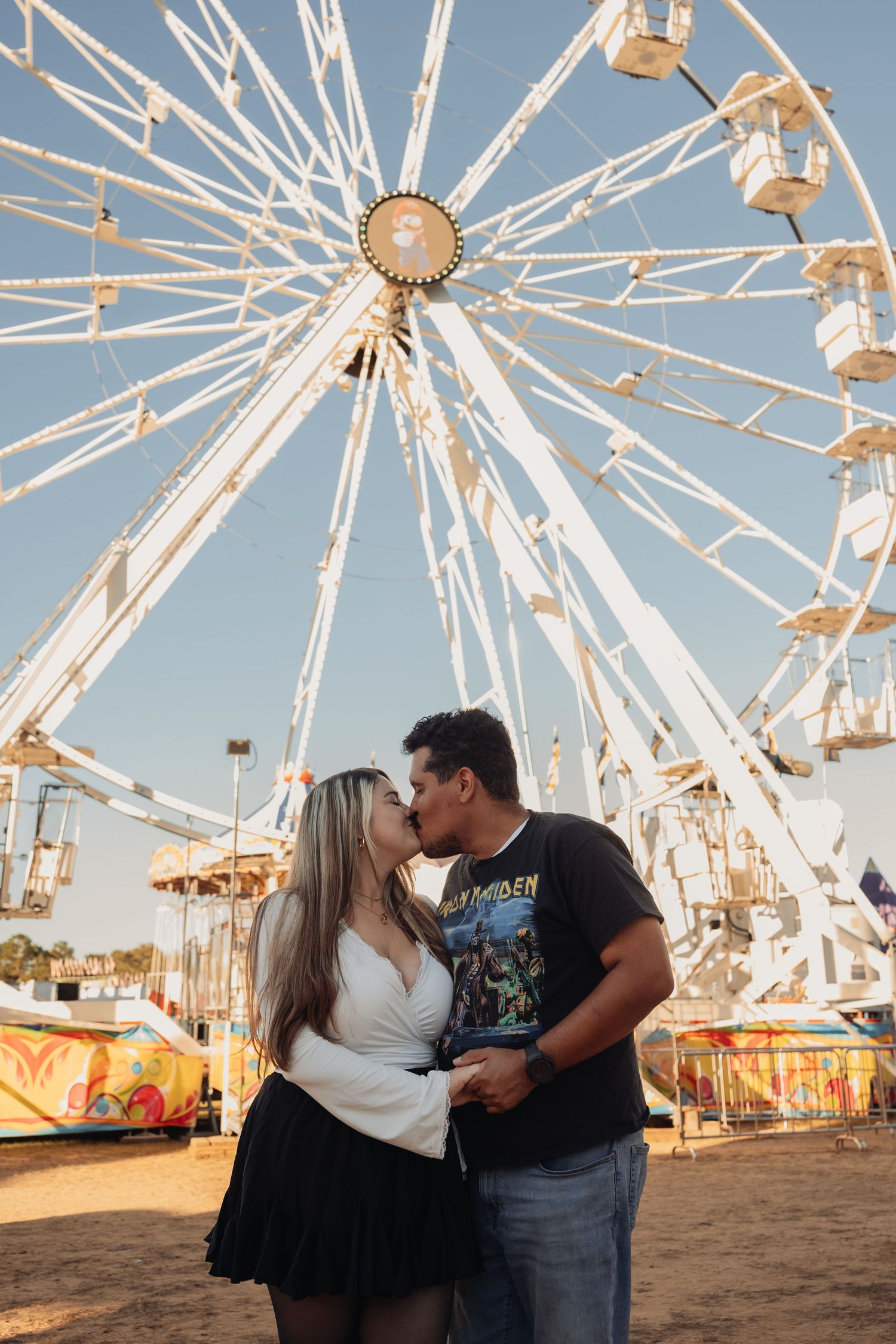 Couple kissing and holding hands in front of a large white Ferris wheel at a sunny carnival fairground under a clear blue sky.