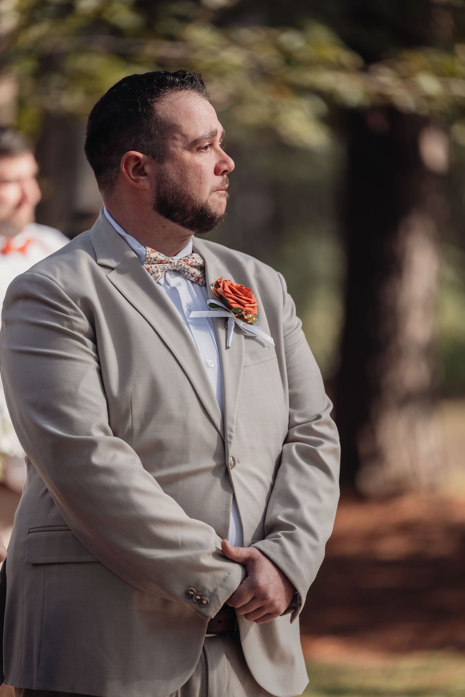 Groom in a tan suit with a floral bow tie and orange boutonniere stands with hands clasped during an outdoor wedding ceremony.