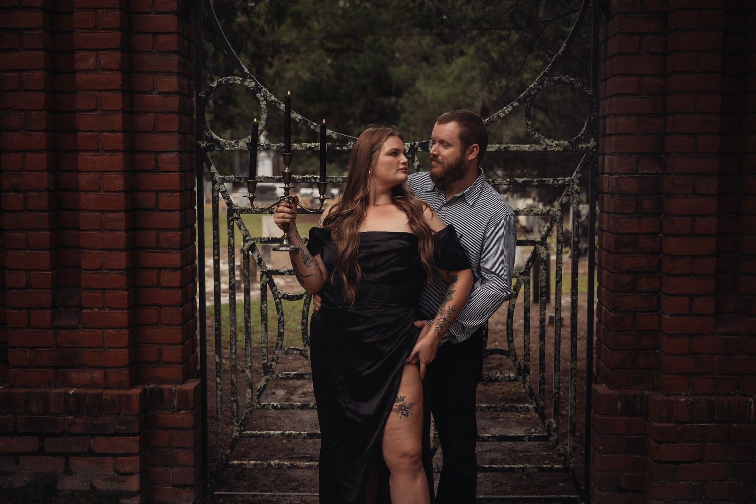 Couple posing romantically at ornate iron cemetery gates, woman in black gown, man in casual shirt with tattoos.