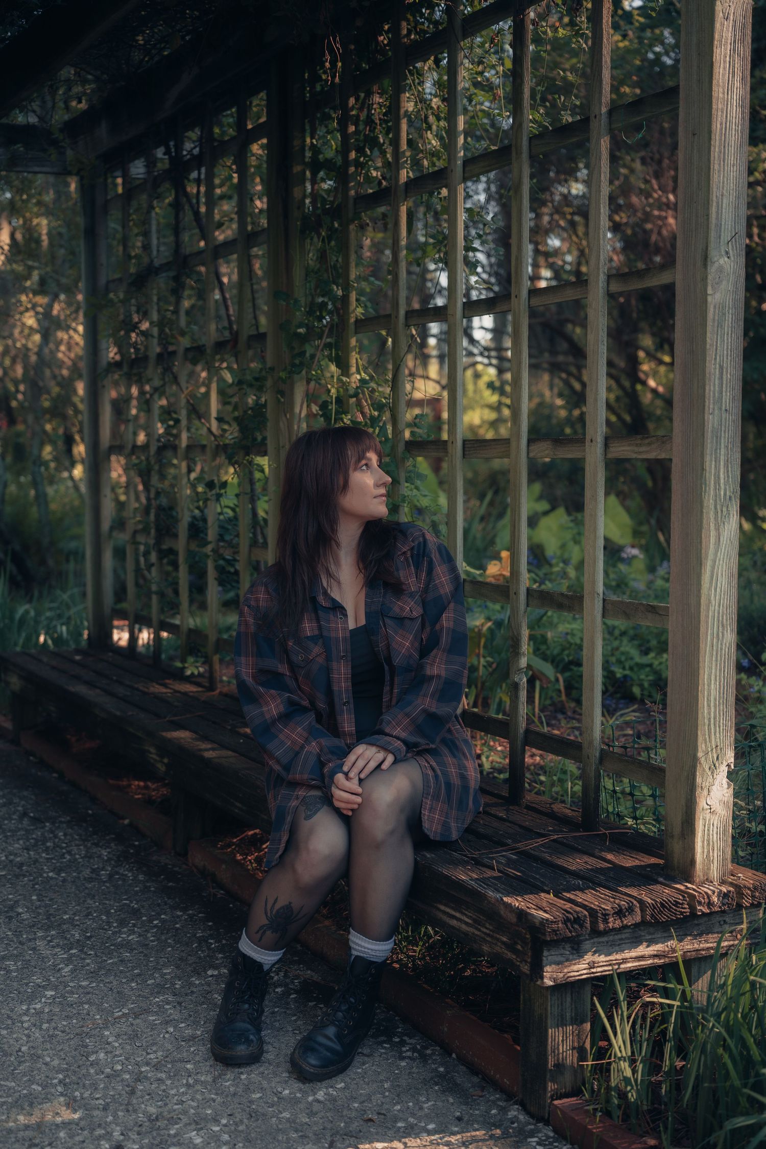 Young woman in plaid shirt sits pensively on wooden bench inside a rustic greenhouse surrounded by lush greenery.