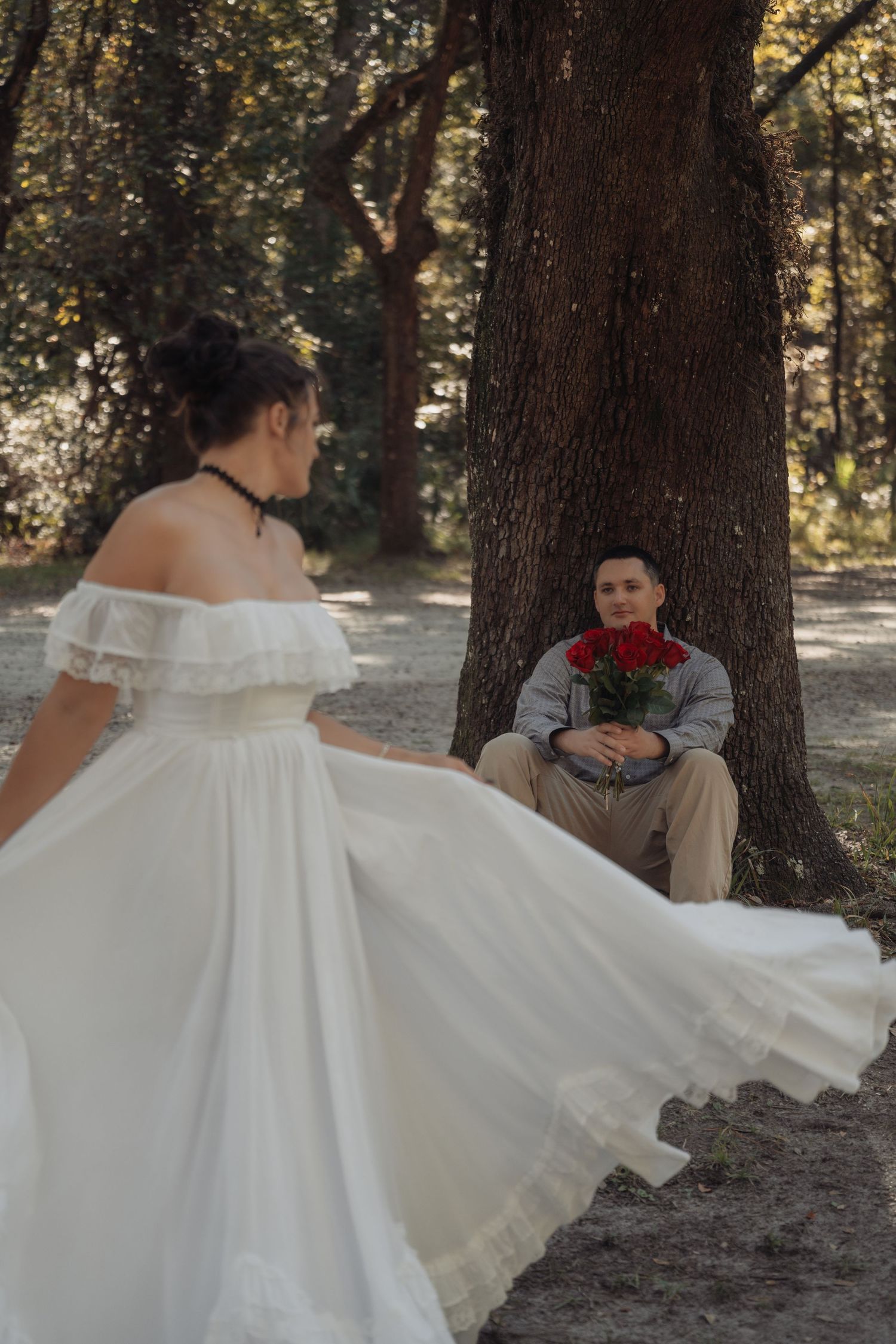 Bride in flowing white off-shoulder gown twirling toward groom holding red roses while leaning against a tree in a forest.