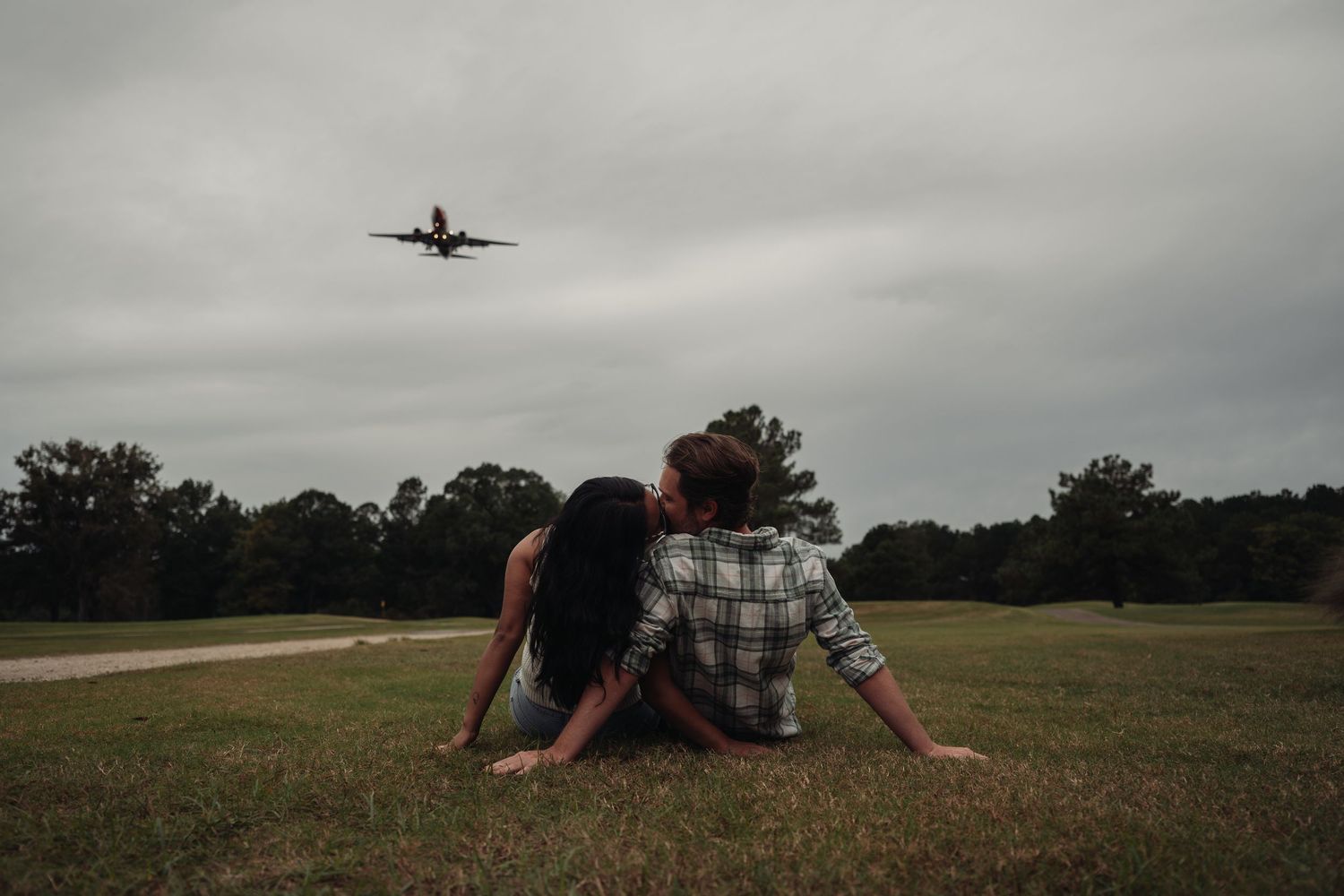 Couple sitting together on grass watching a plane fly overhead on a cloudy day.