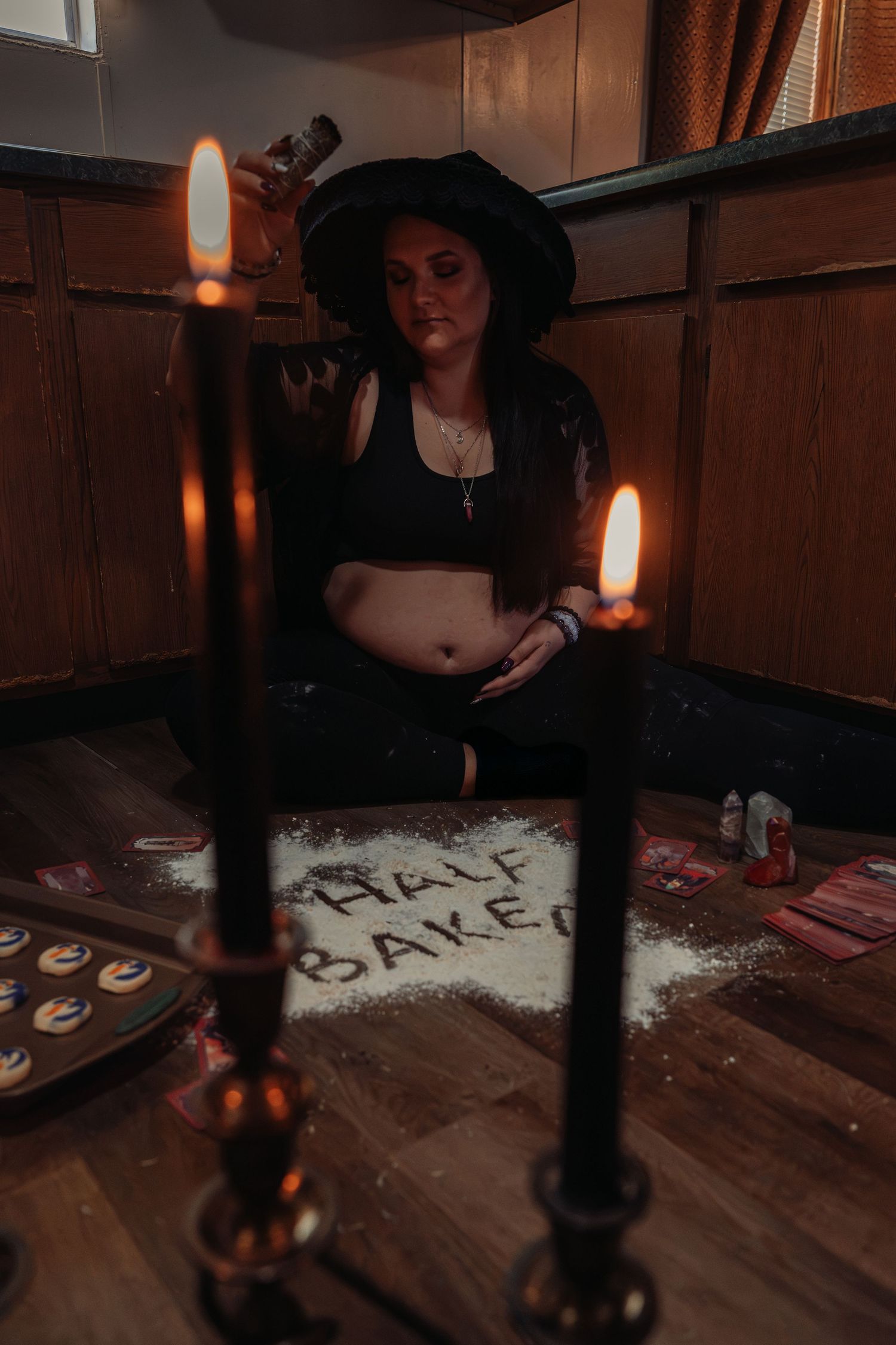 Witch-themed woman sitting on floor surrounded by candles and Half Baked flour writing in dim room.