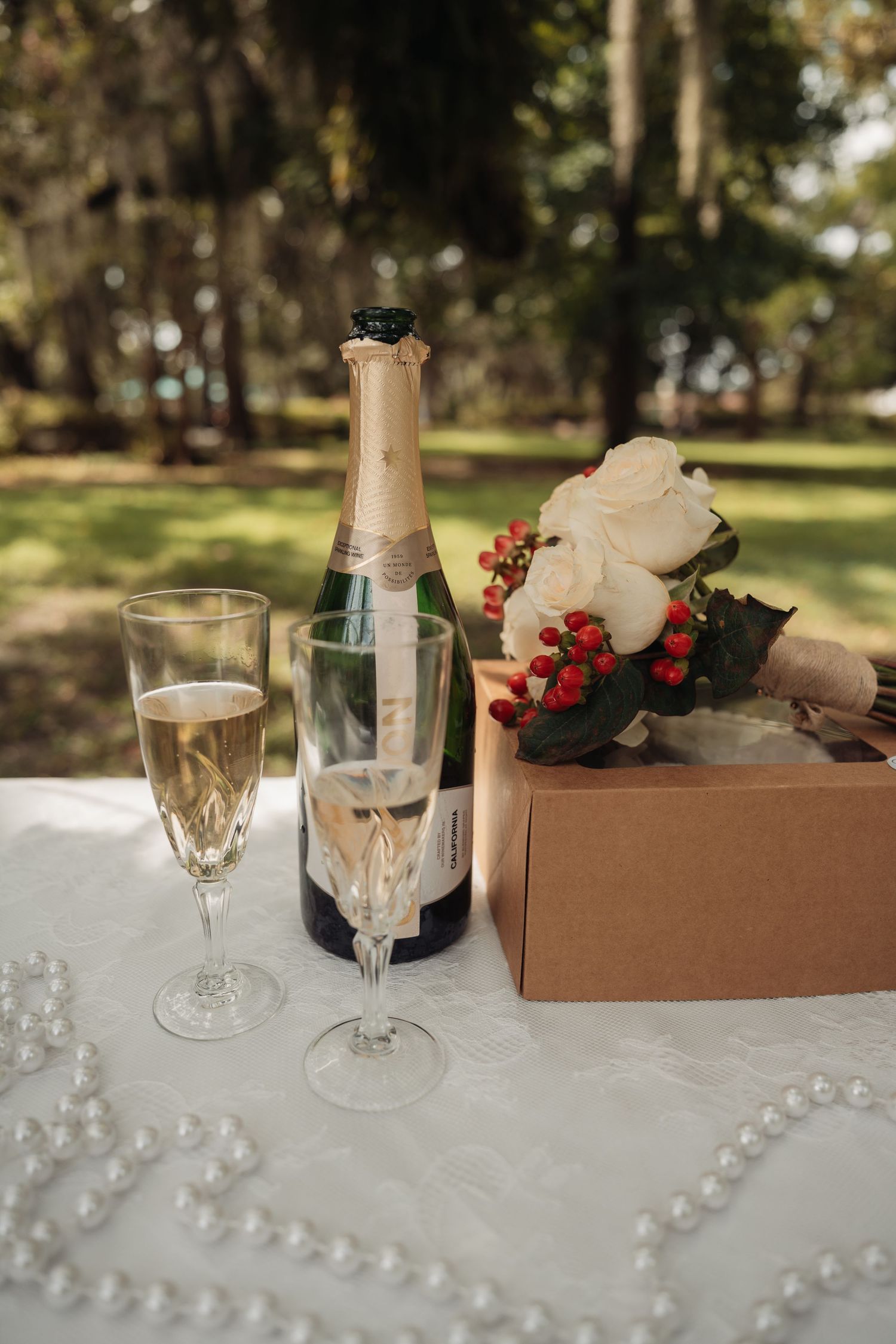 A wedding bouquet of white roses and red berries sits beside champagne glasses and bottle on a table.