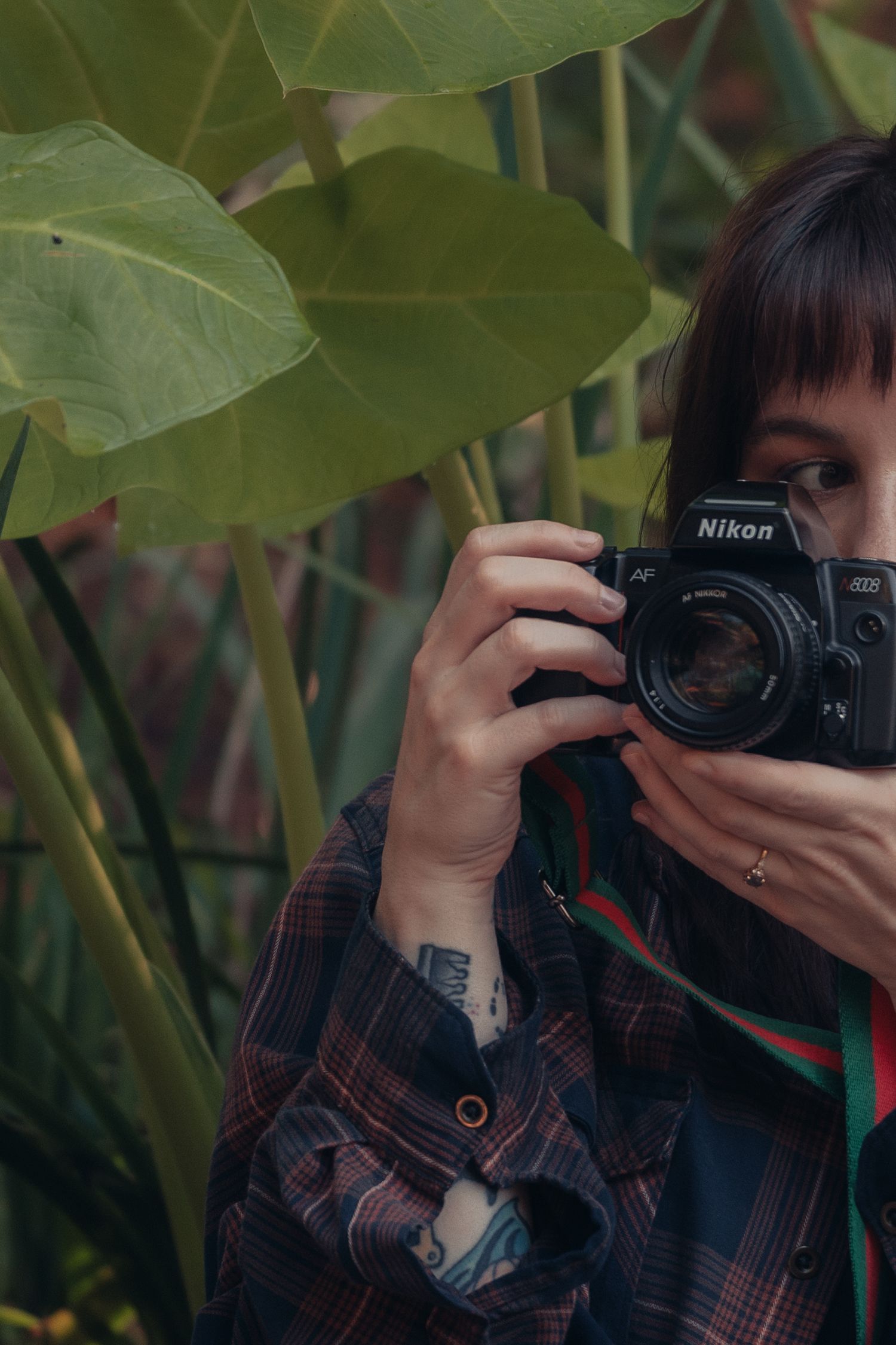 Woman with tattoos holding a Nikon film camera up to her face surrounded by large tropical green leaves.
