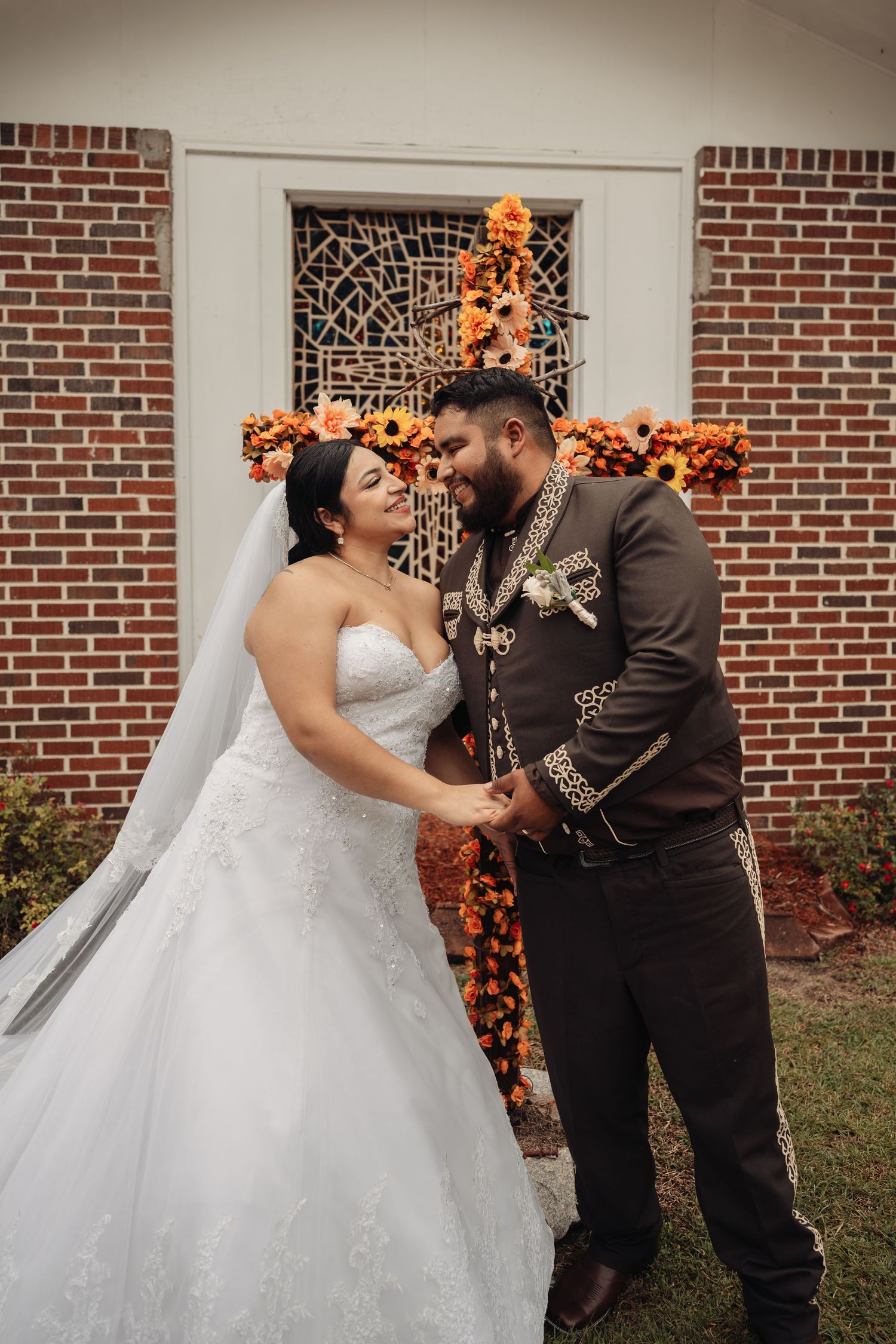 Bride and groom holding hands at outdoor wedding ceremony with orange floral cross backdrop against brick church wall.