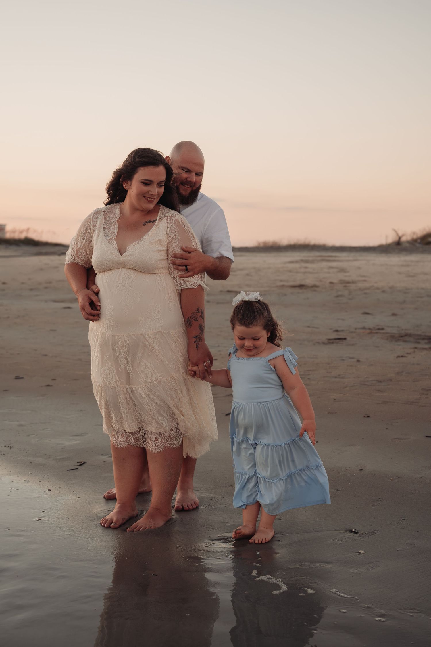 A smiling couple stands embracing on a wet sandy beach at sunset while holding hands with their young daughter in a blue dress.