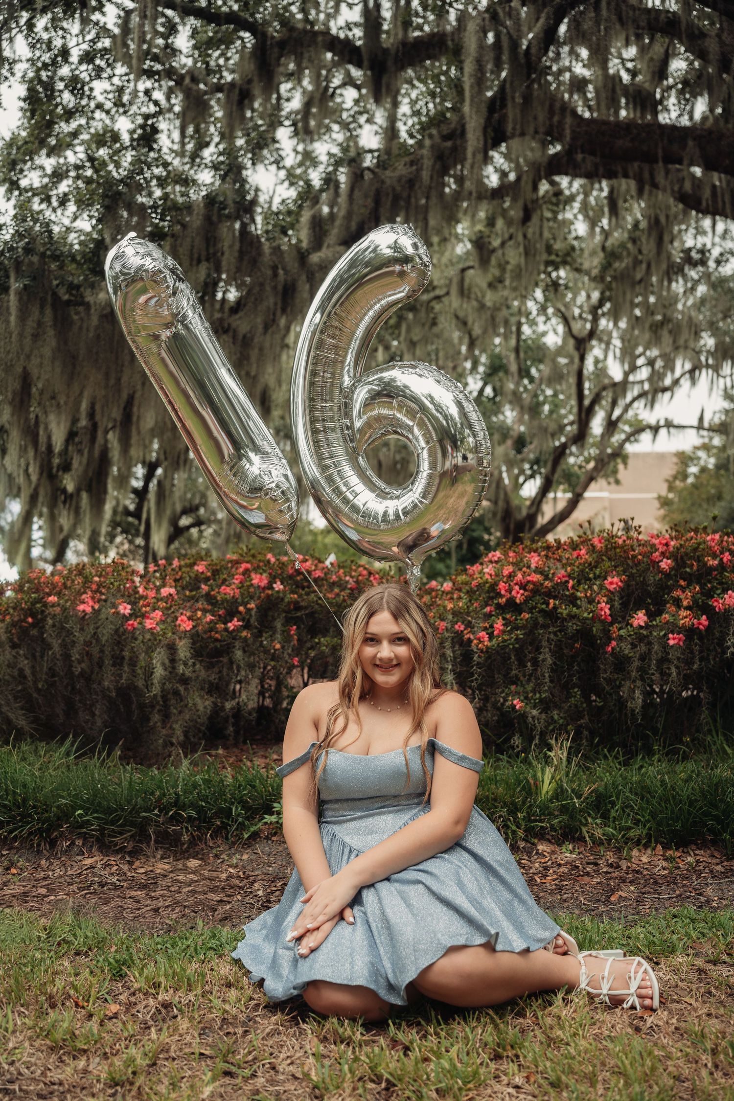 A teenage girl in a blue dress sits on grass holding silver number 16 balloons for her birthday celebration outdoors.