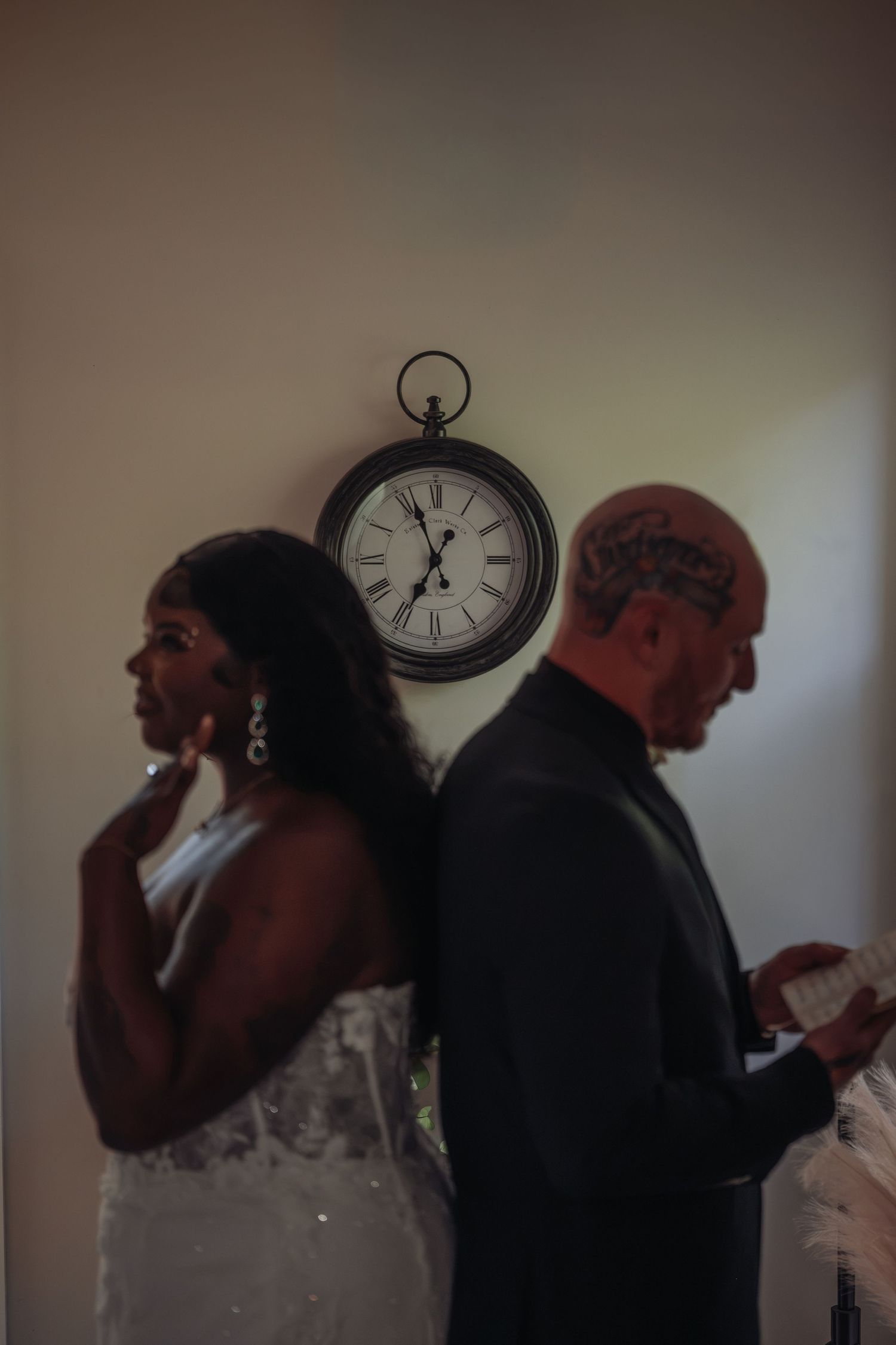 Bride and groom stand back-to-back reading vows with vintage clock on wall behind them.
