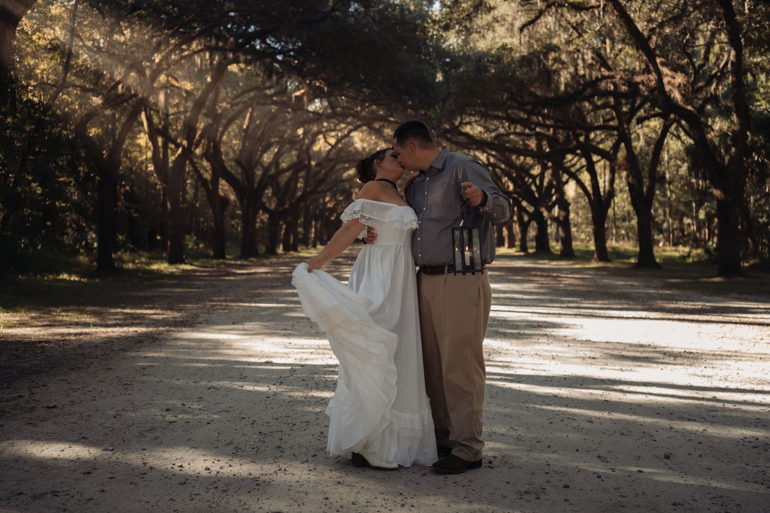 Couple sharing a romantic dip kiss on a sunlit tree-lined path, woman in elegant white off-shoulder wedding gown.
