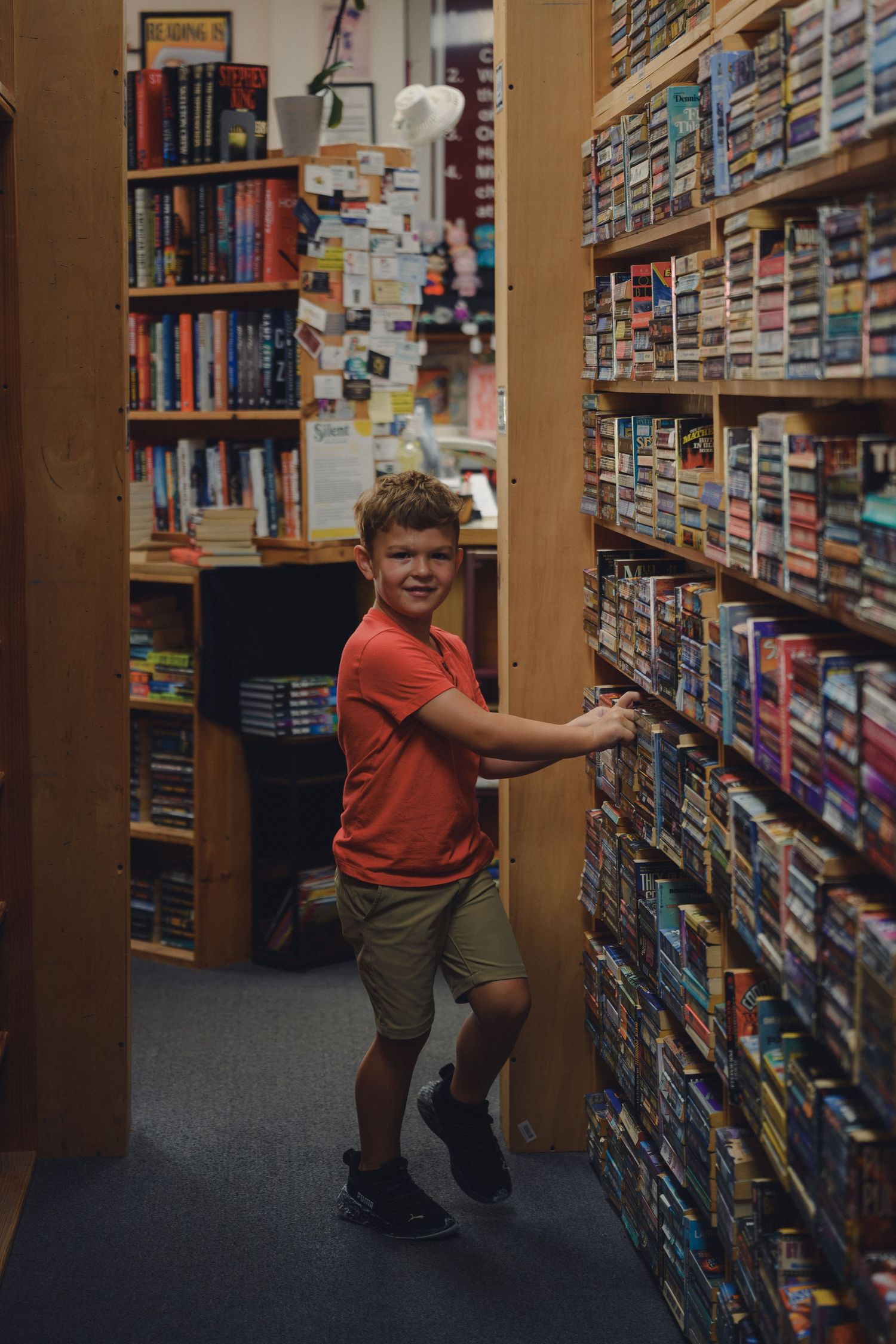 A smiling boy in a red shirt browses shelves of books and videos in a cozy, well-stocked bookstore aisle.