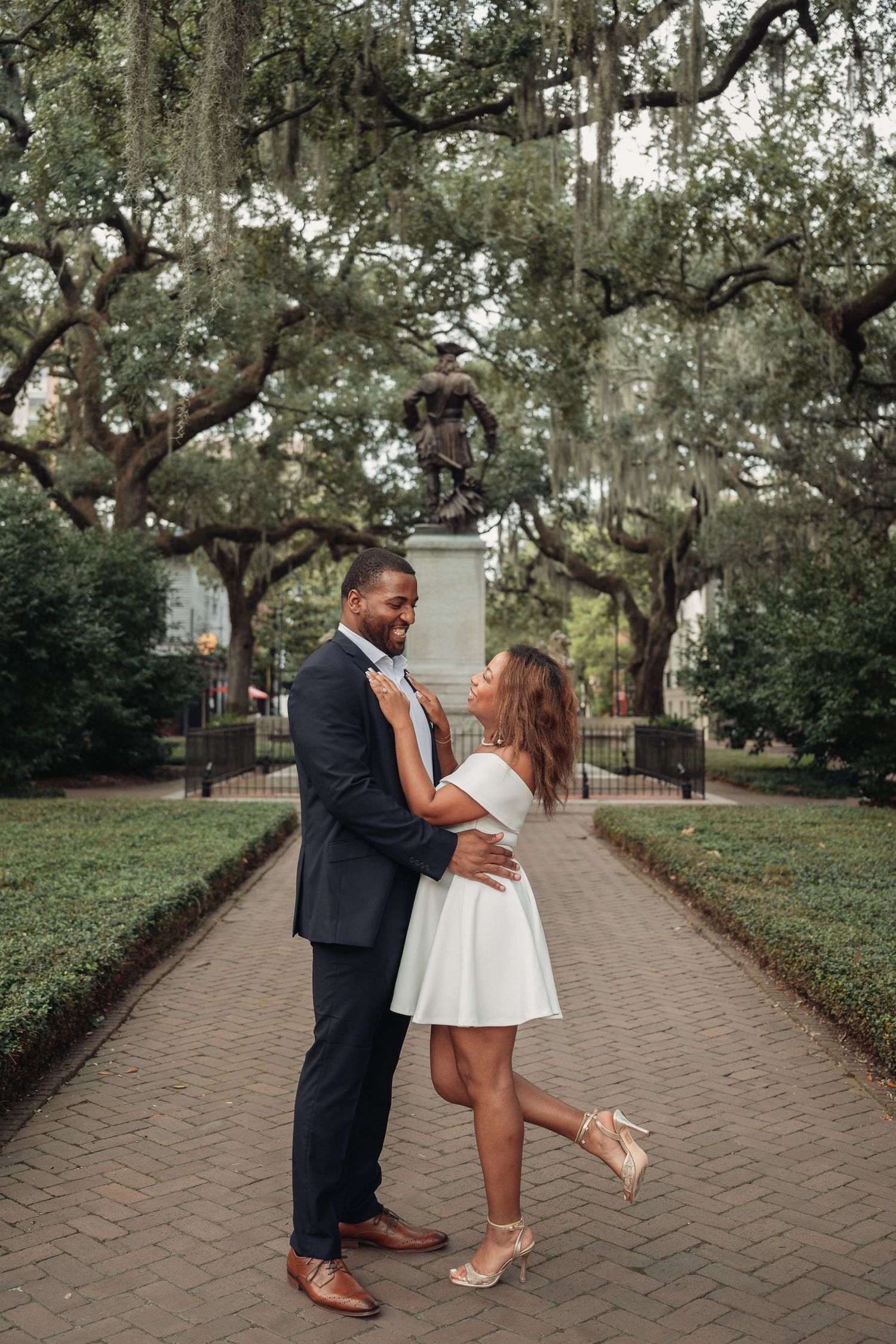 Joyful couple dancing on brick path in Savannah square, woman lifting leg in white dress and heels, man in navy suit.