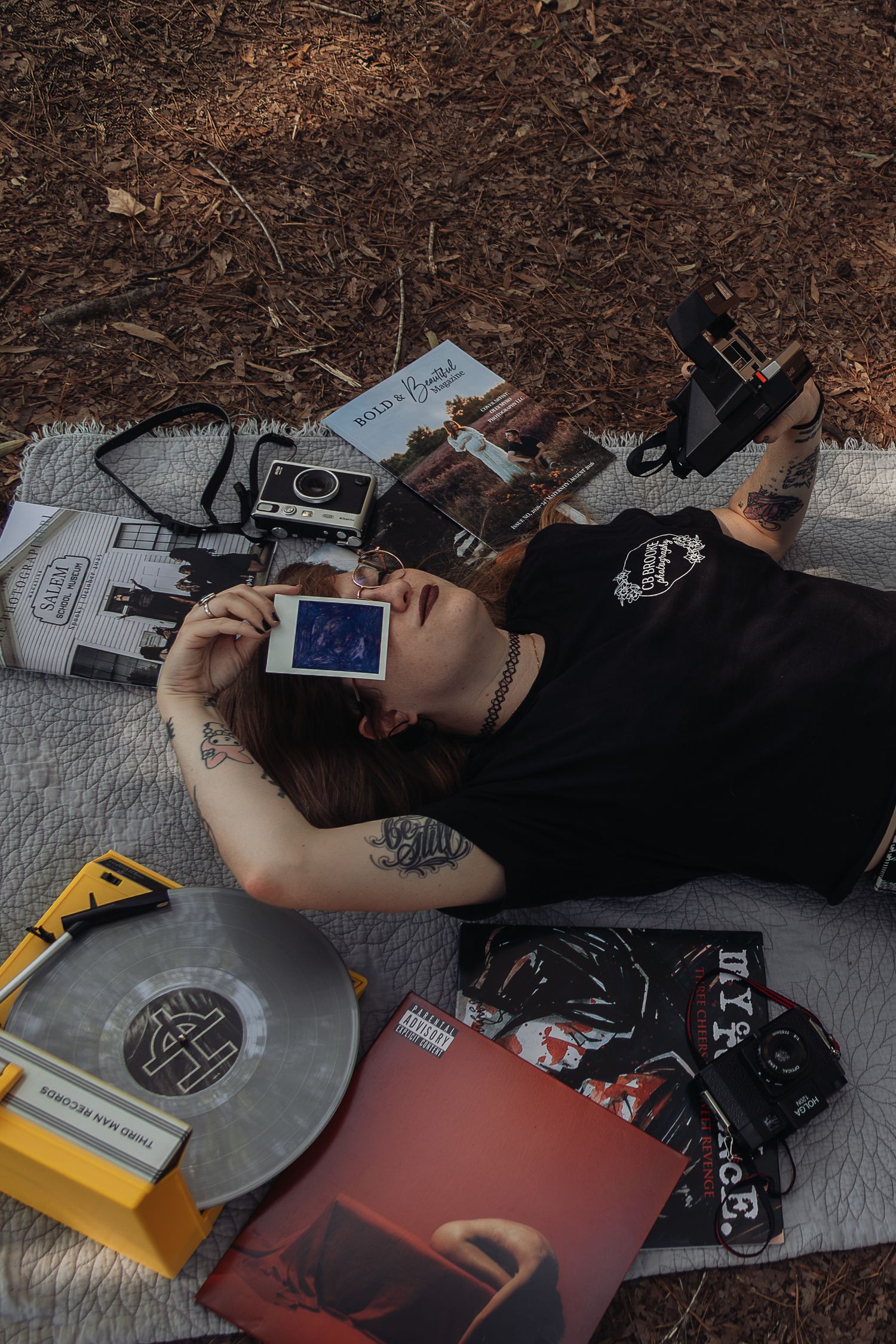 Woman lying on blanket holding Polaroid photo up to face surrounded by vinyl records and cameras.