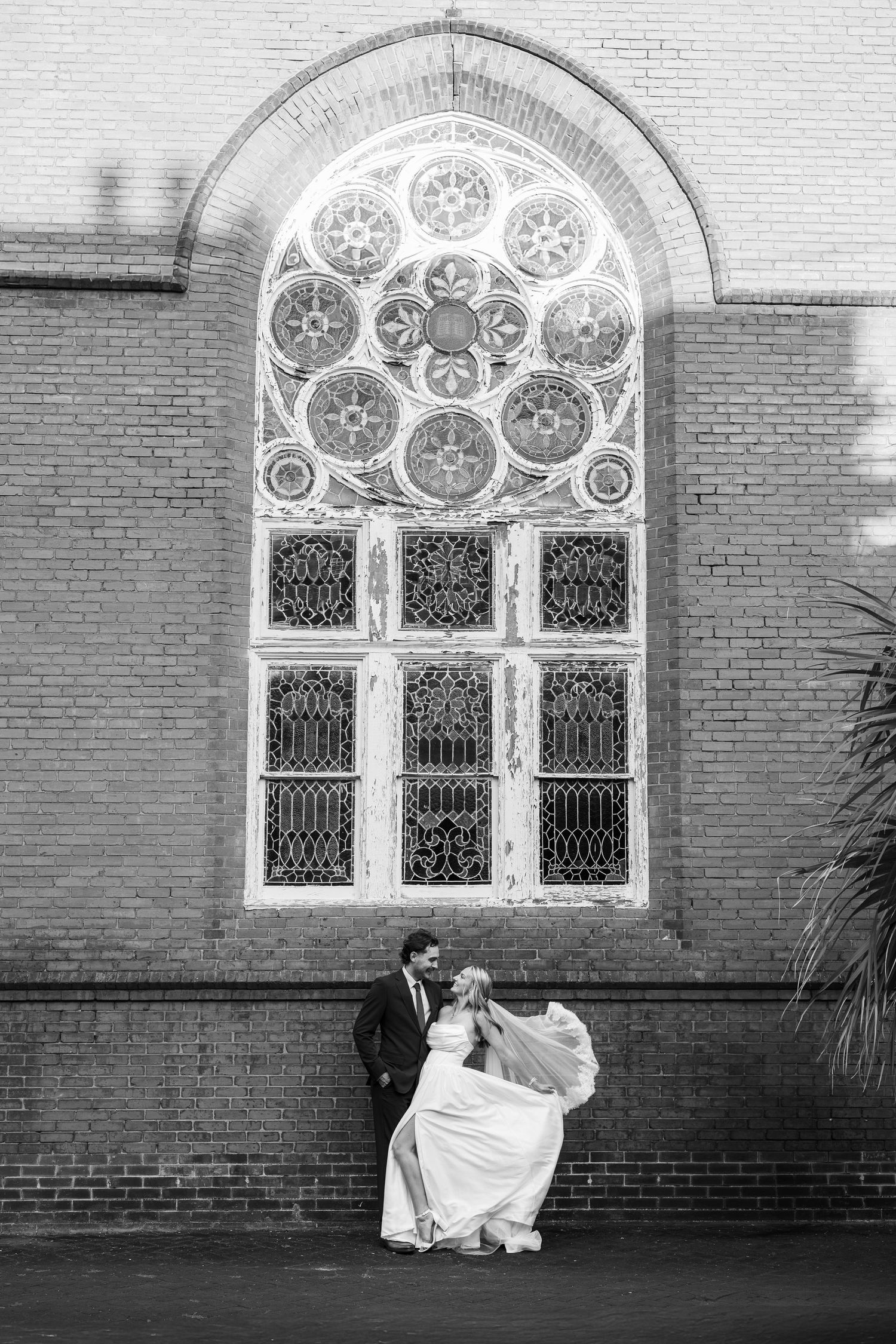 Black and white photo of bride and groom dancing before a large ornate stained glass window.