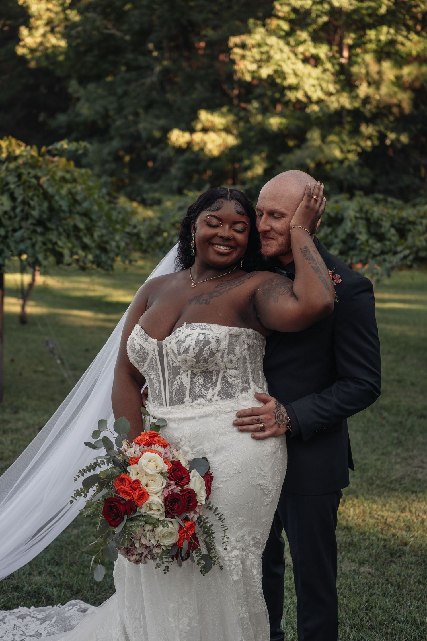 Smiling bride in lace gown with red bouquet embraced by bald groom outdoors.