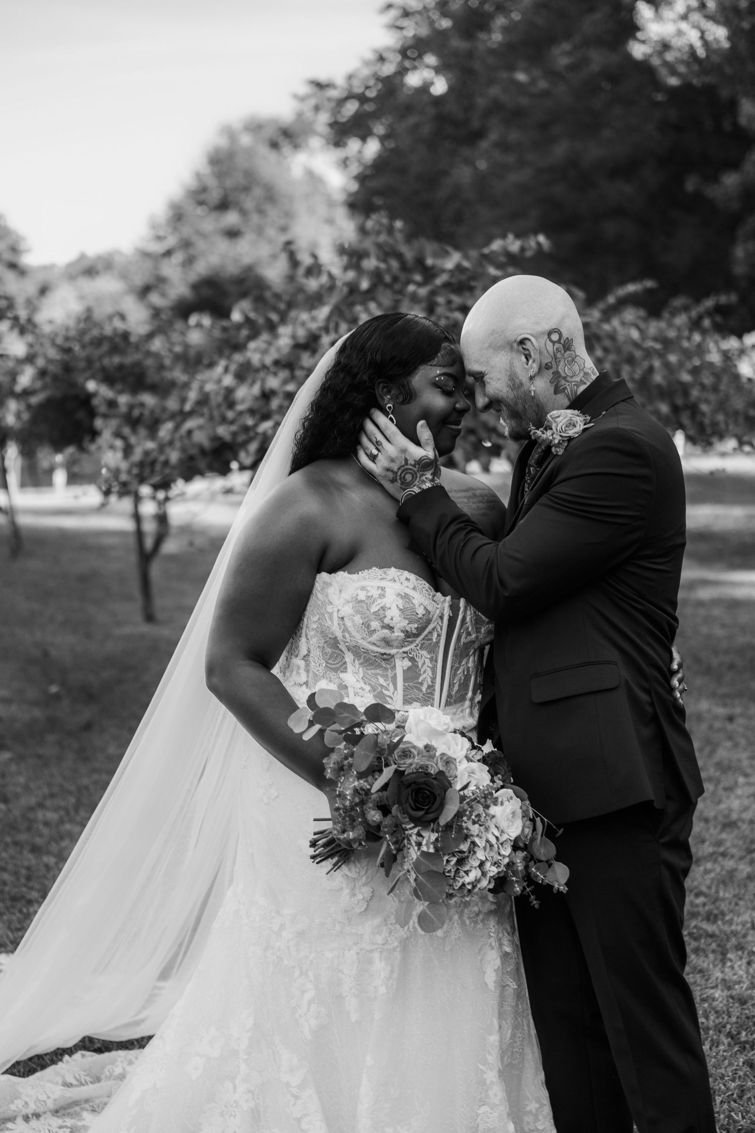 Black and white romantic portrait of bride and tattooed groom nearly kissing in a garden.
