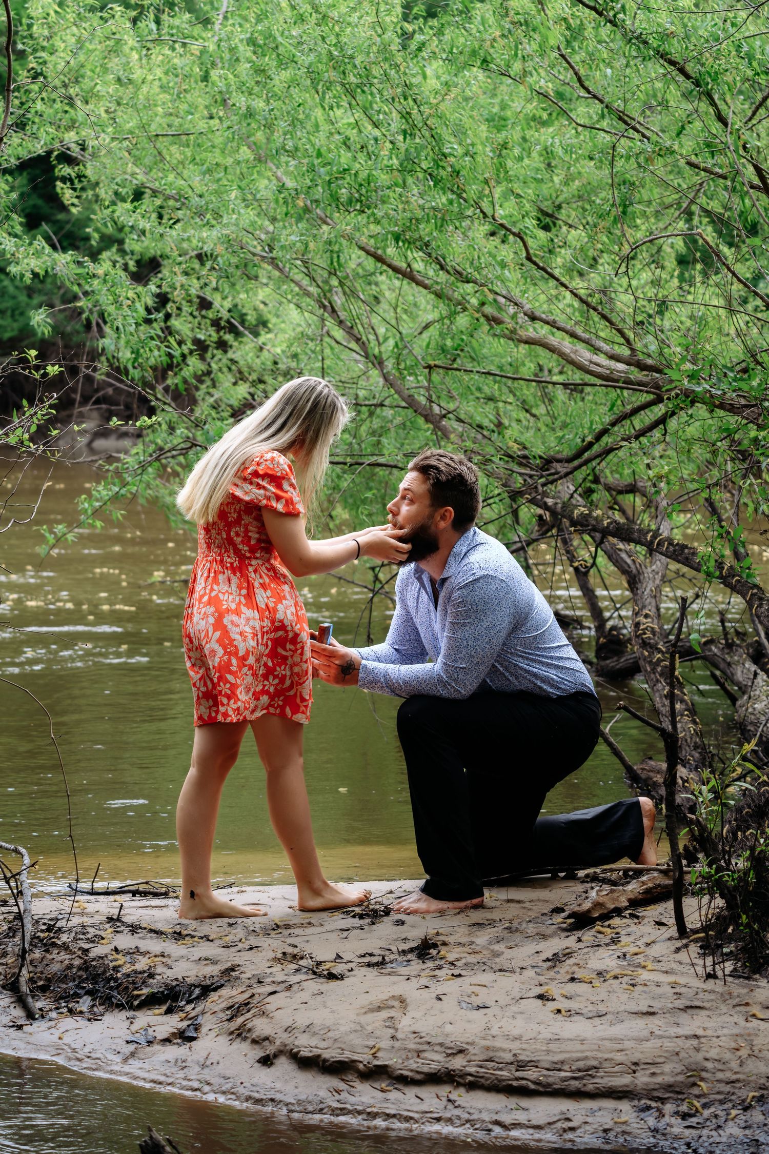 Man proposing on one knee to woman in red dress by a river surrounded by green trees.