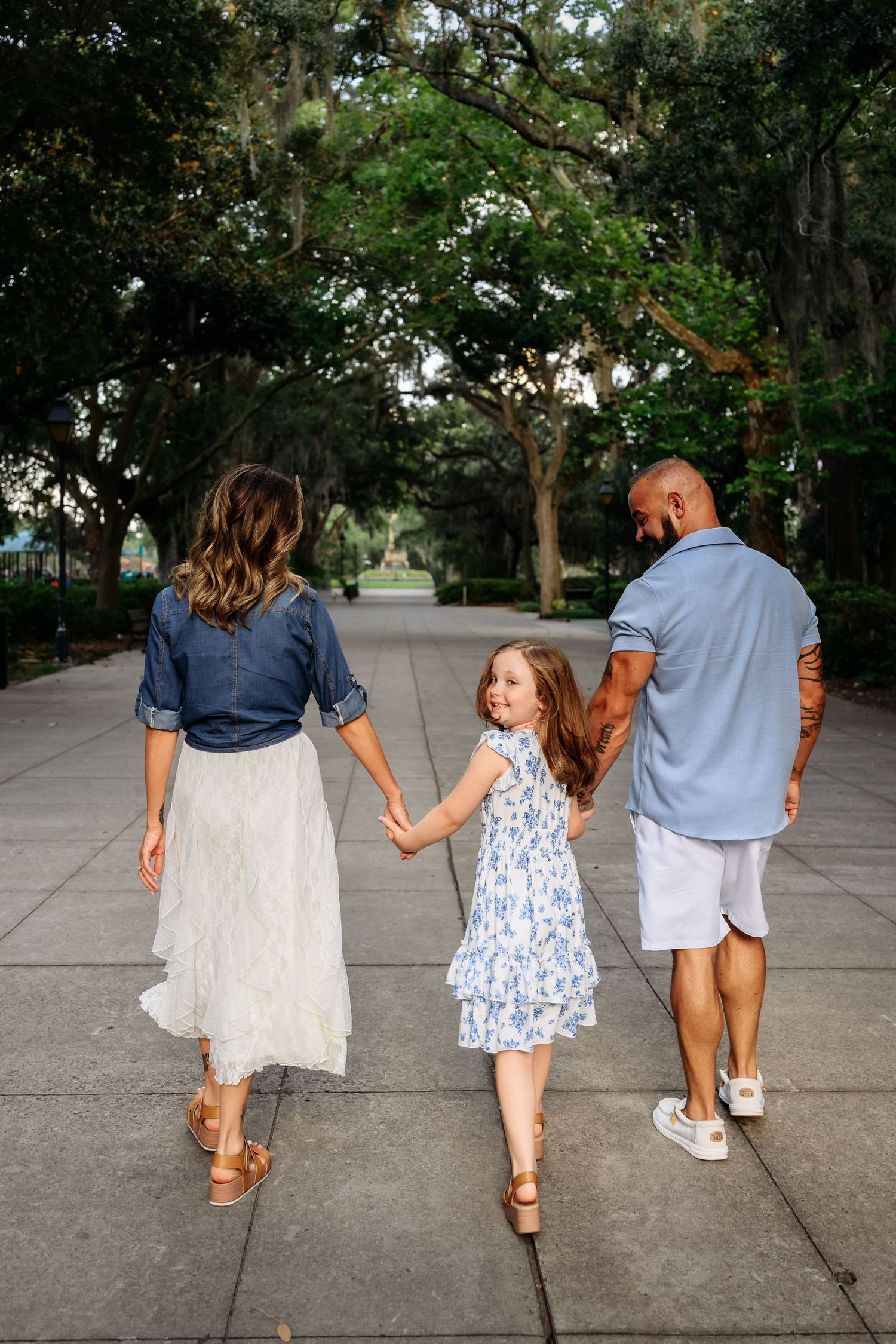 Family of three walking hand-in-hand down a tree-lined path, wearing coordinated blue and white outfits.