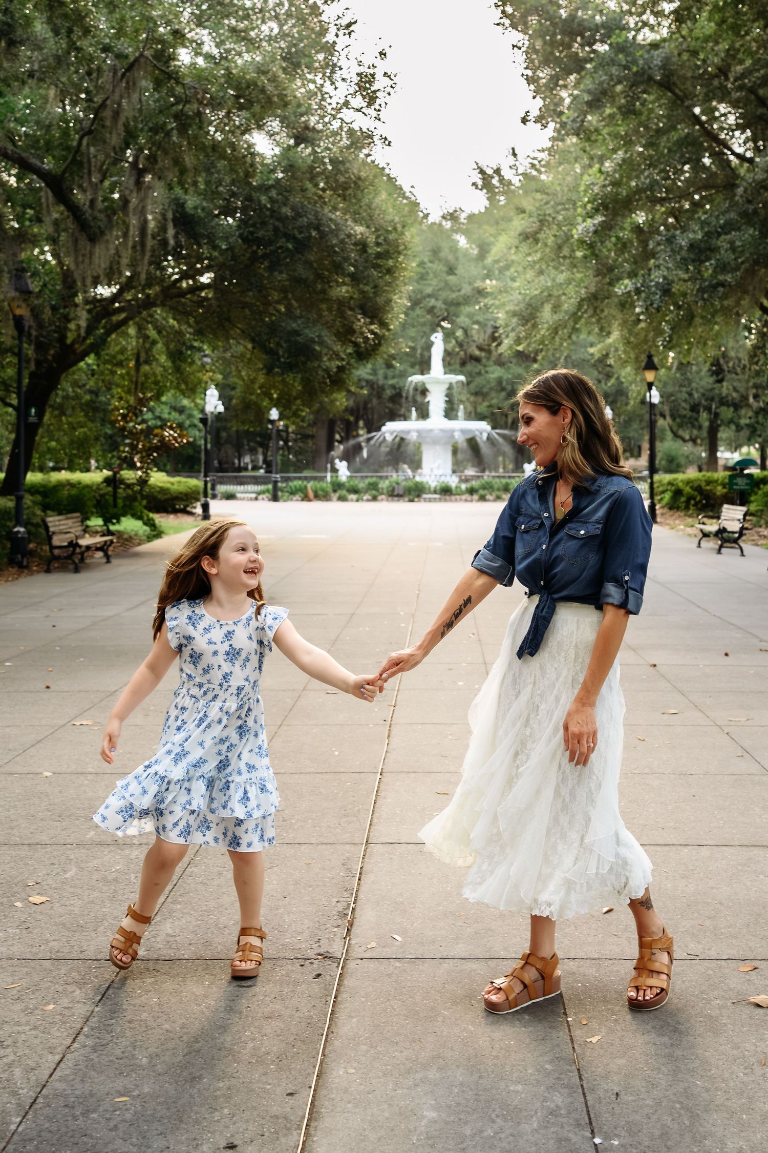 A woman and young girl hold hands and dance in a park with a fountain, both wearing dresses and sandals.