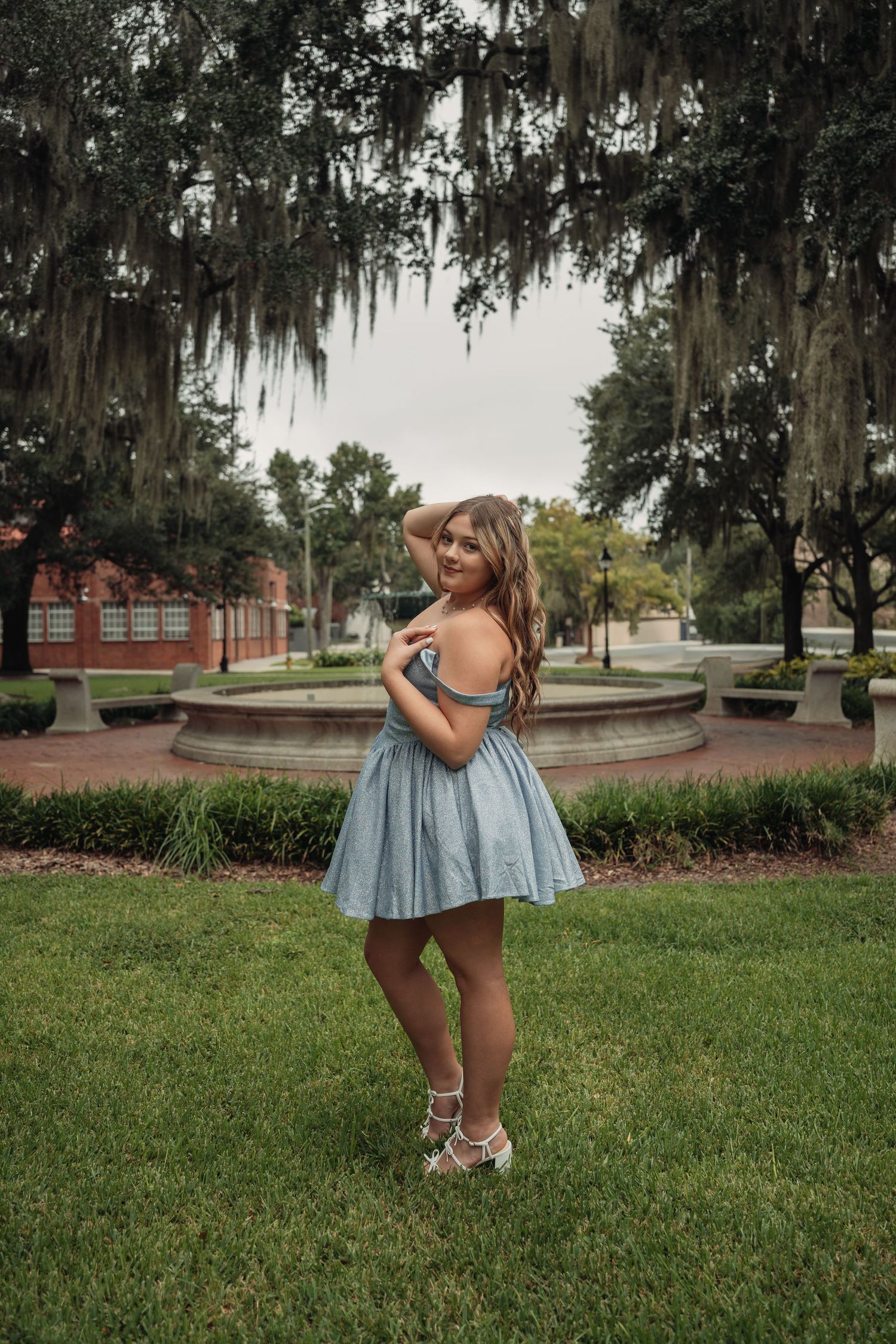 Young woman in blue off-shoulder dress poses gracefully in a lush park with Spanish moss-draped trees and fountain.