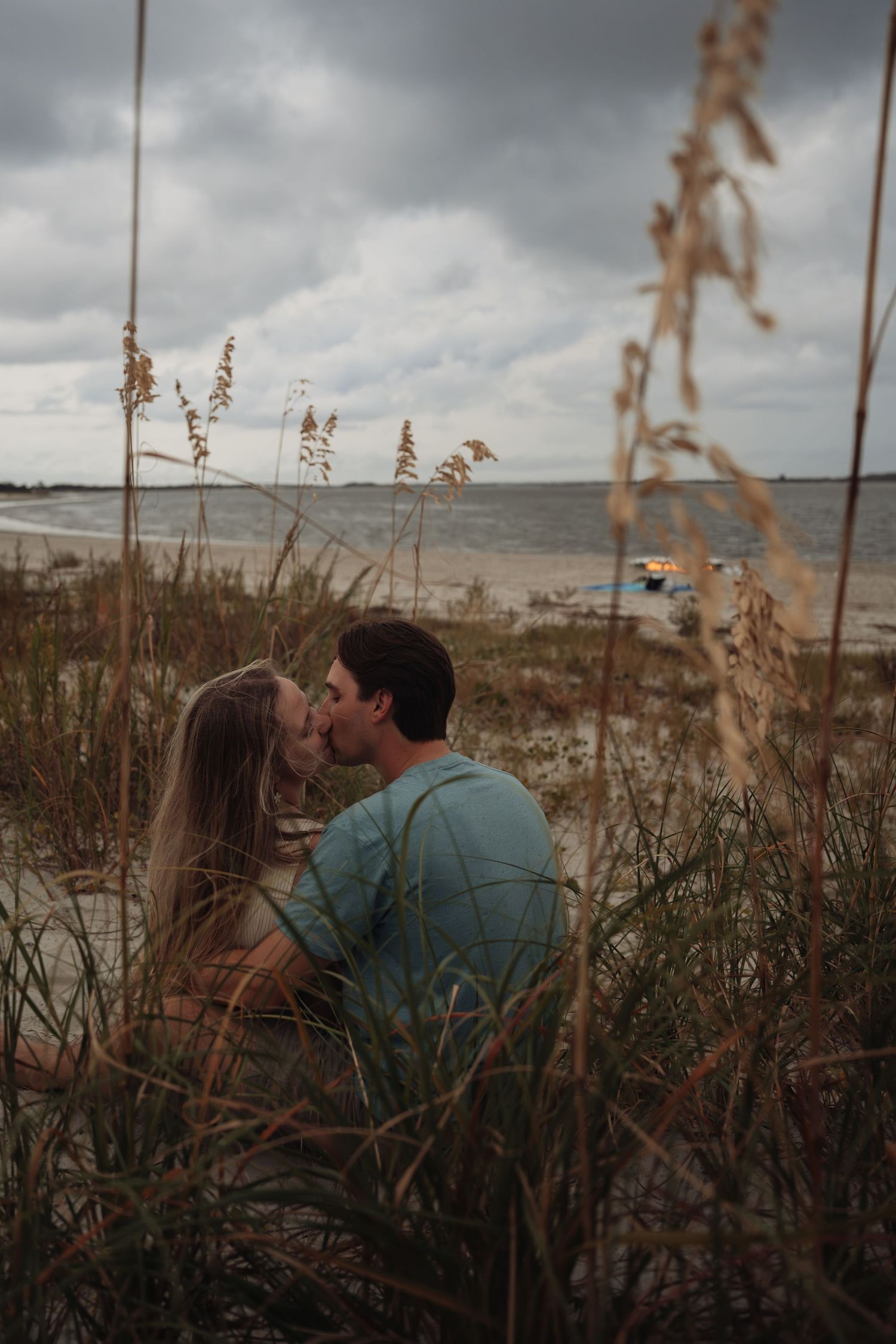 Couple kissing on a sandy beach framed by sea oats grass under cloudy skies.