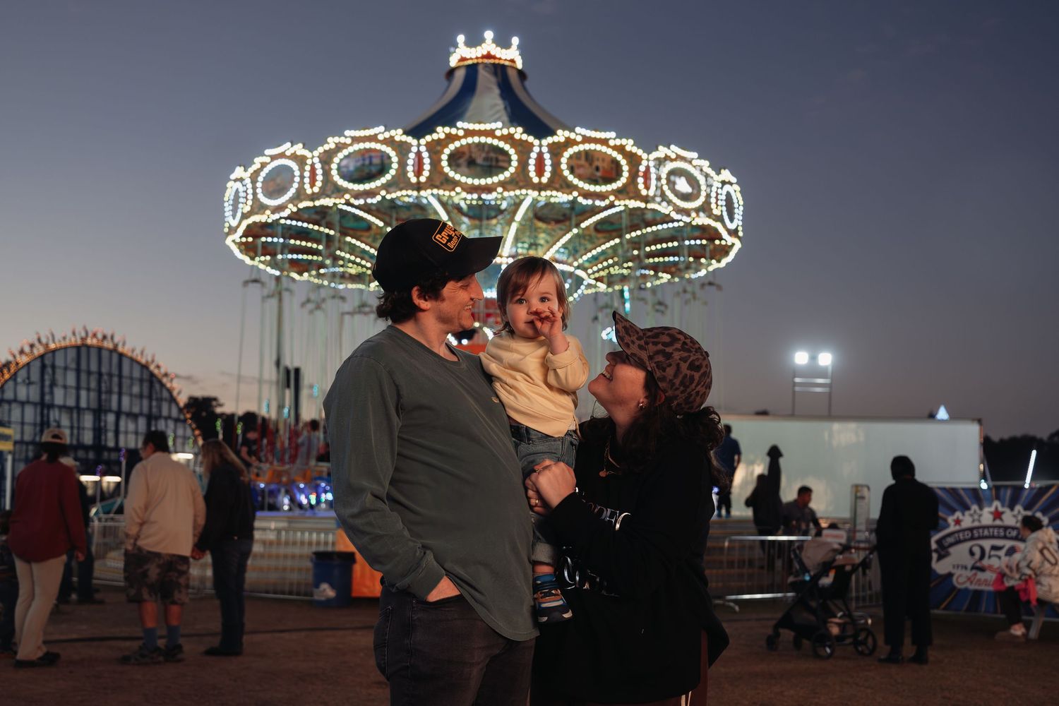 A smiling couple holds a toddler at a carnival at dusk, with a brightly lit swing carousel ride and fairgoers in the background.