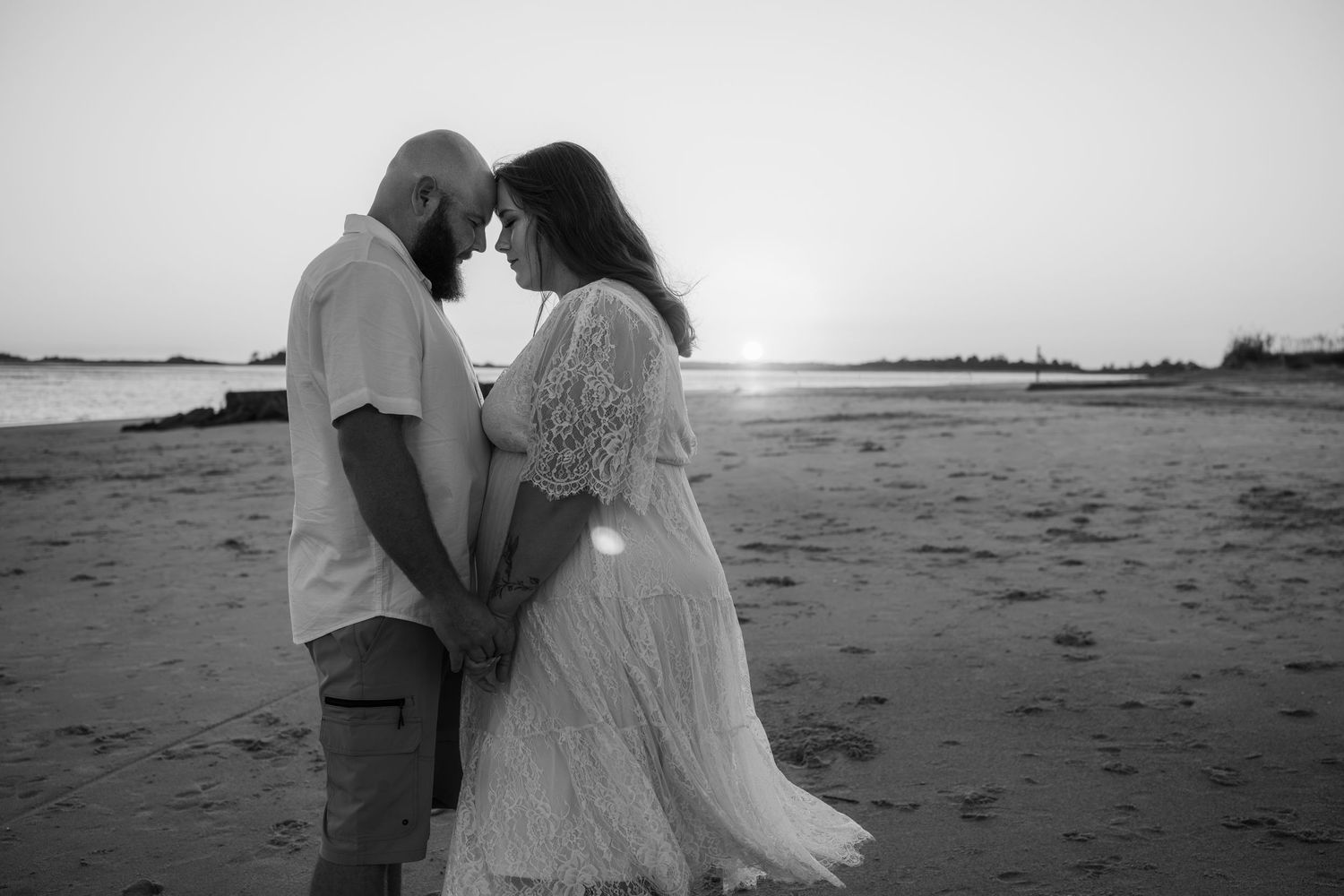 Black and white photo of a couple standing forehead to forehead and holding hands on a sandy beach at sunset.