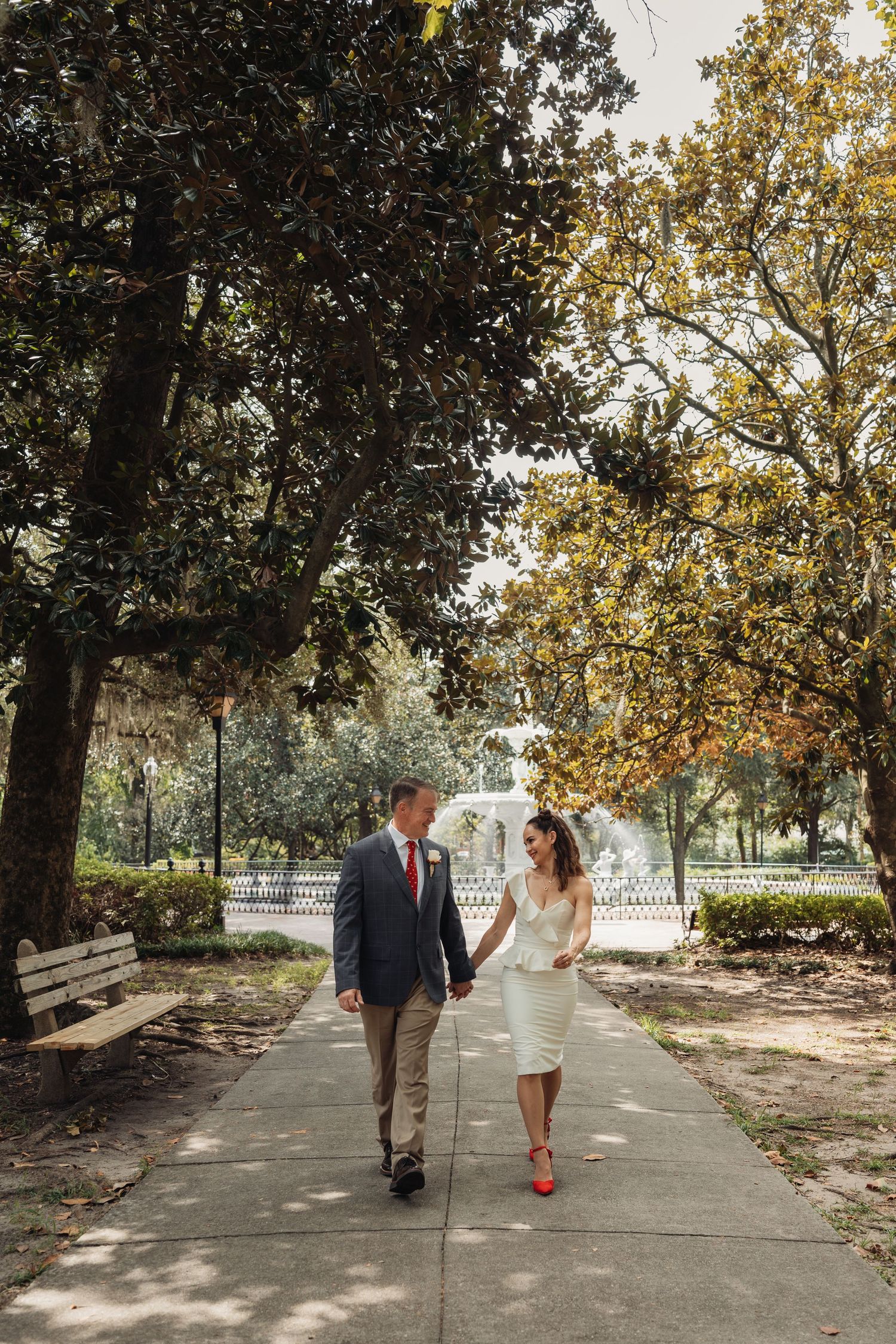 Newlyweds walk hand-in-hand along a tree-lined park path with a fountain visible in the background.