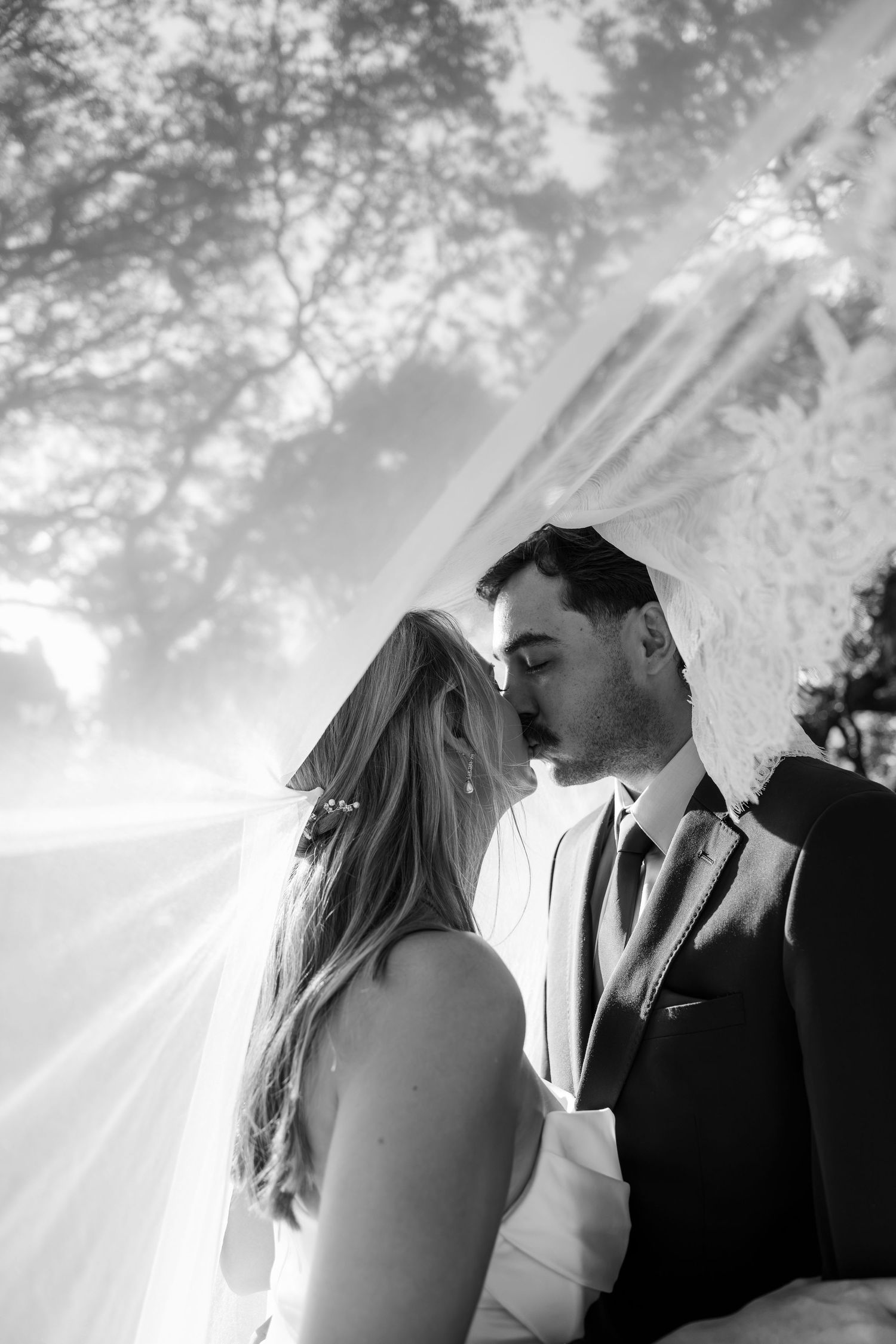 Black and white wedding photo of bride and groom sharing an intimate moment under a flowing veil outdoors.
