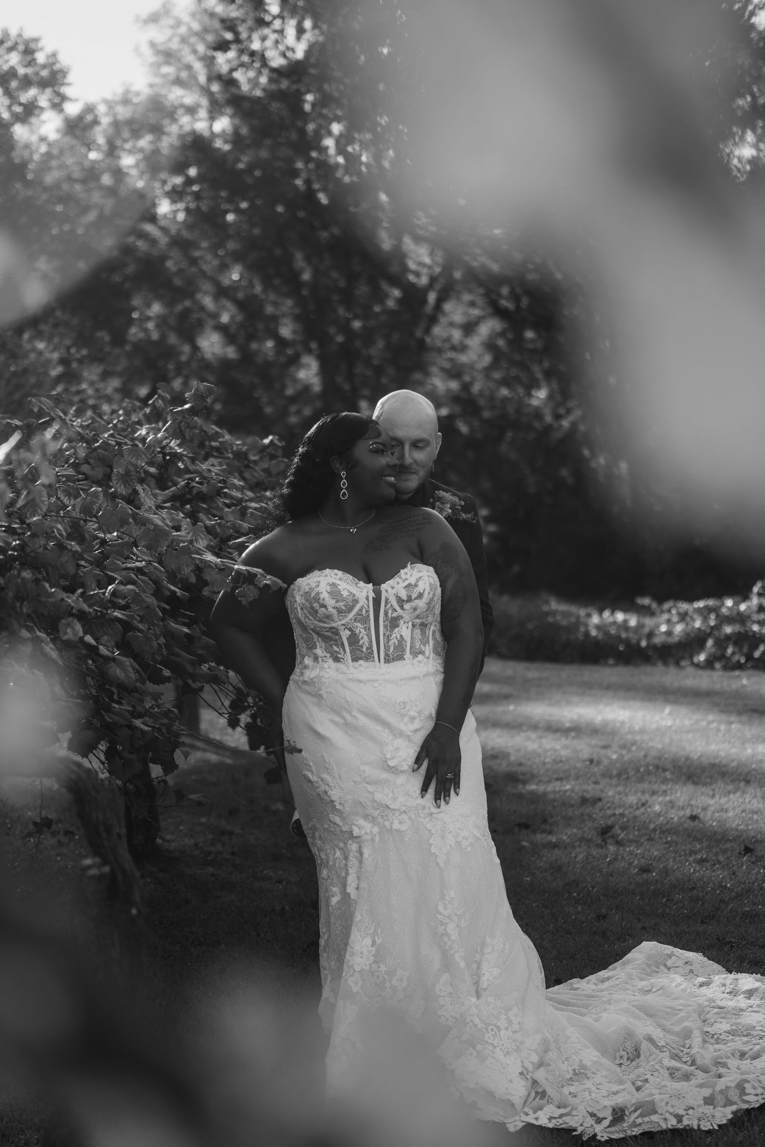 Black and white portrait of bride in lace gown and groom kissing her cheek in a garden.