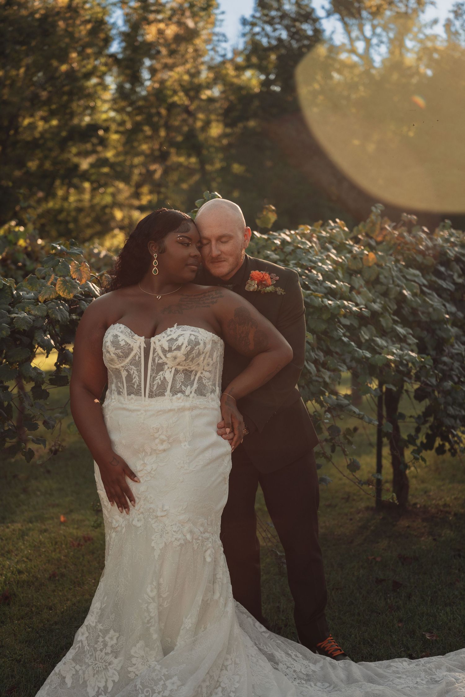 Bride in lace mermaid gown and groom share golden hour moment in vineyard setting.