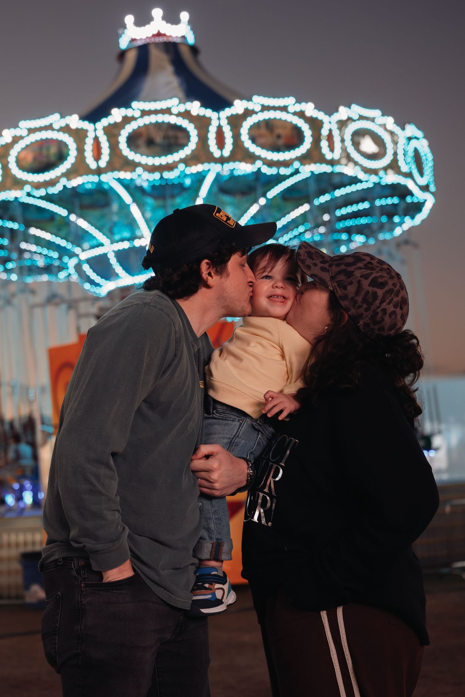 Two parents kiss a smiling toddler on the cheeks in front of a glowing, illuminated carousel at an evening amusement park.