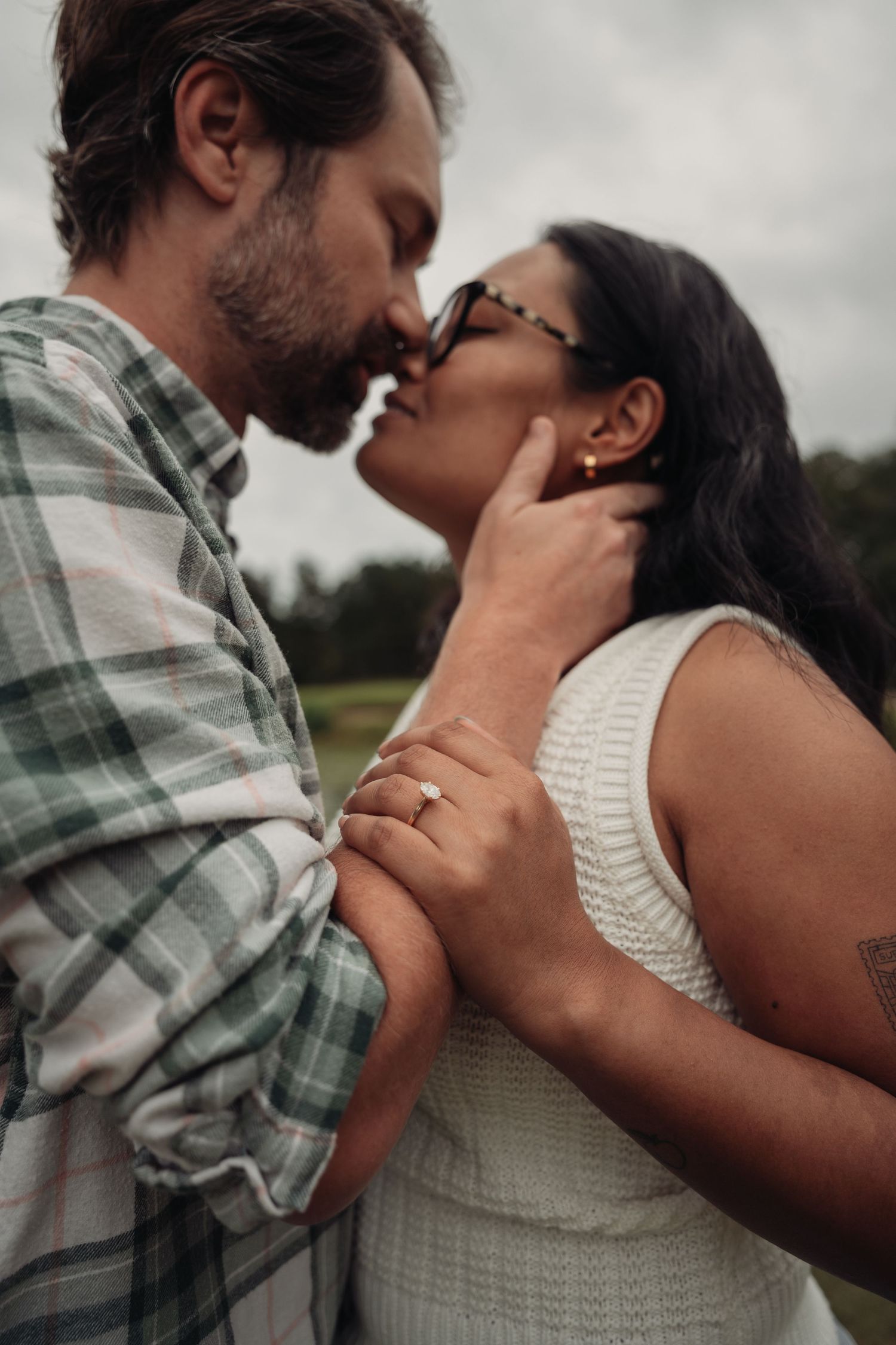 A couple shares a romantic almost-kiss outdoors, the woman wearing glasses and an engagement ring, dressed in white.