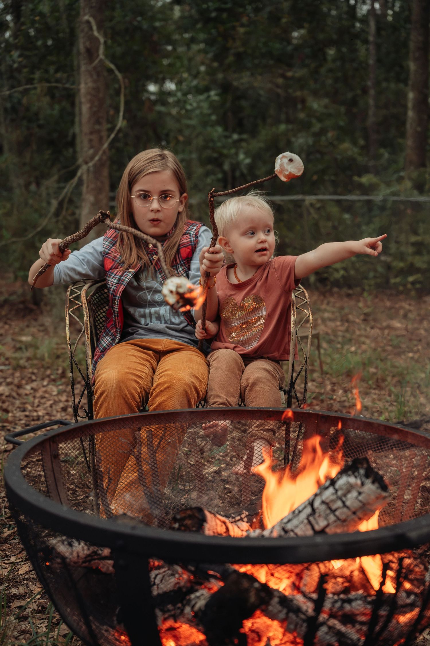 Two children roasting marshmallows over a campfire in the woods, sitting in a metal chair outdoors.