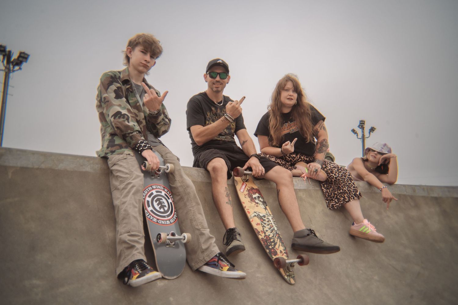 Four skaters sit on the edge of a concrete skatepark ramp under a hazy sky, two holding skateboards and one flashing a hand sign.