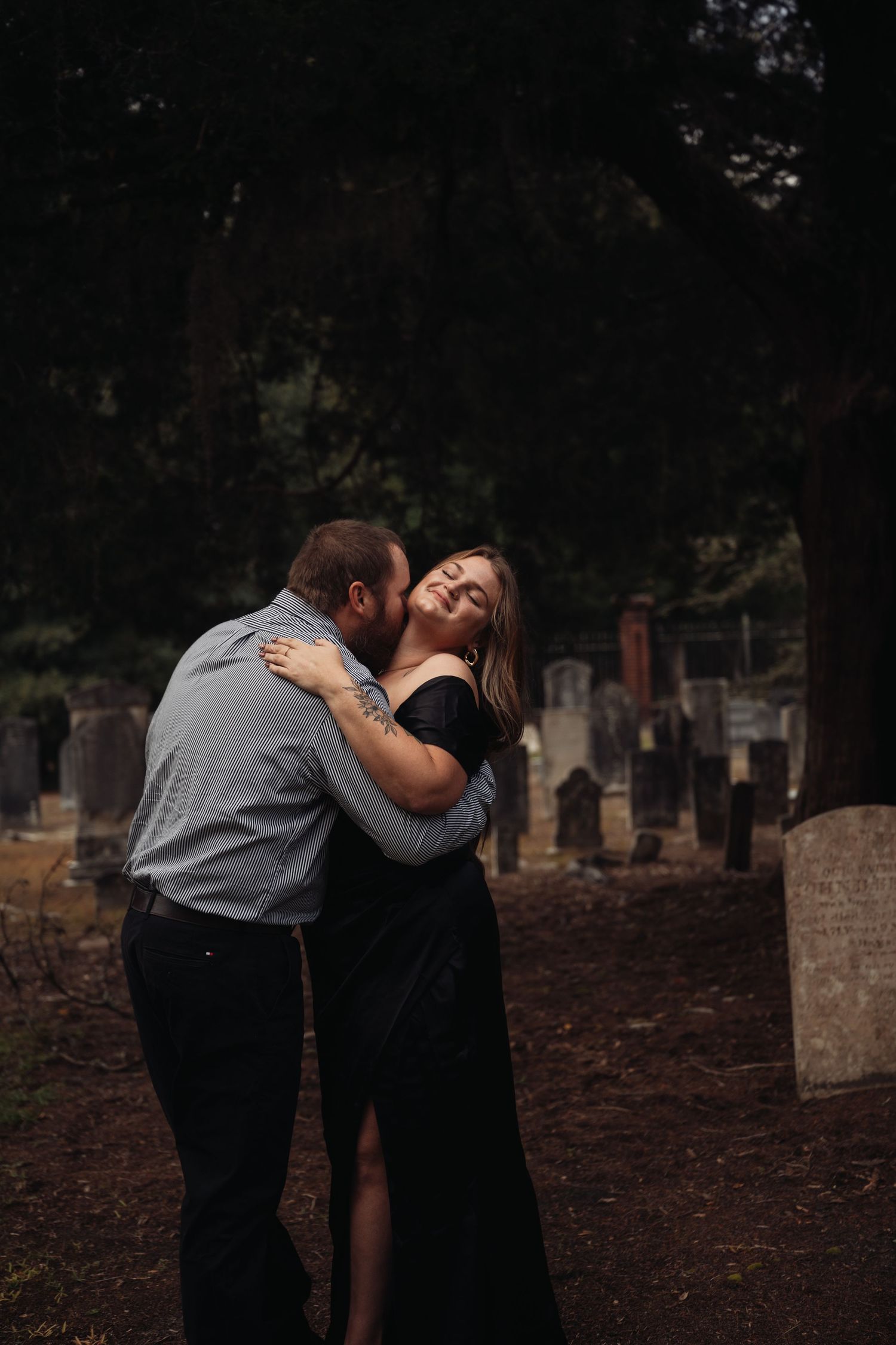Couple embracing romantically in a cemetery, woman in black dress laughing joyfully as man kisses her cheek.