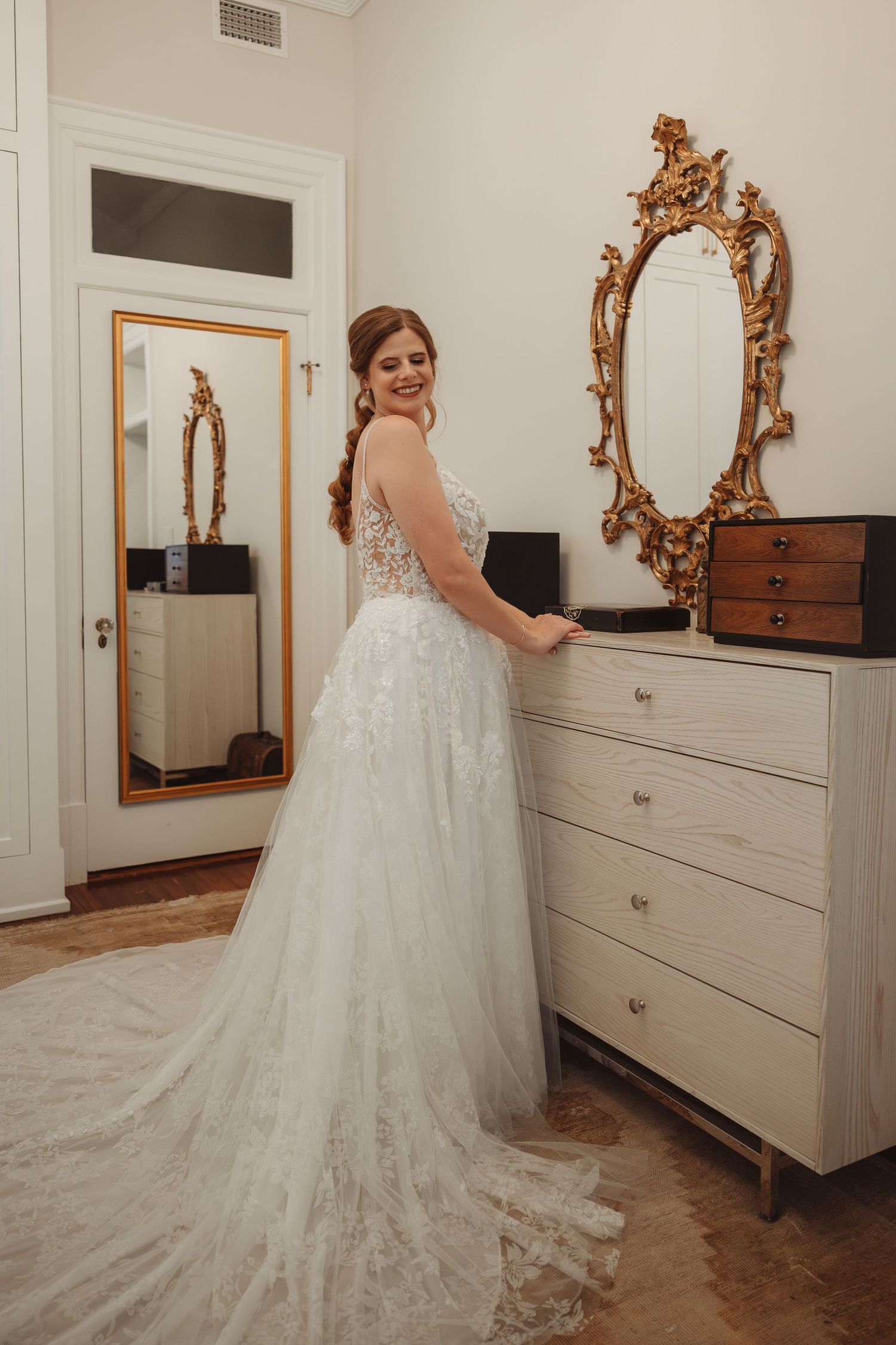 Bride in a flowing lace wedding gown stands by a white dresser with ornate gold mirror in an elegant room.