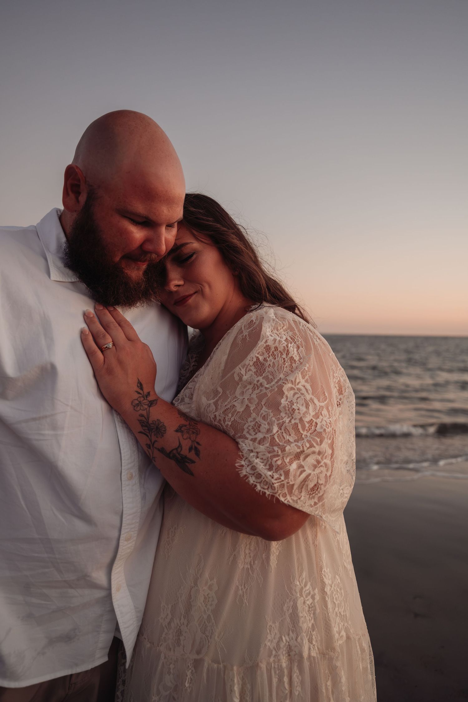 Couple embracing on a beach at sunset, the woman in a cream lace dress resting her head on a bearded man's chest by the ocean.