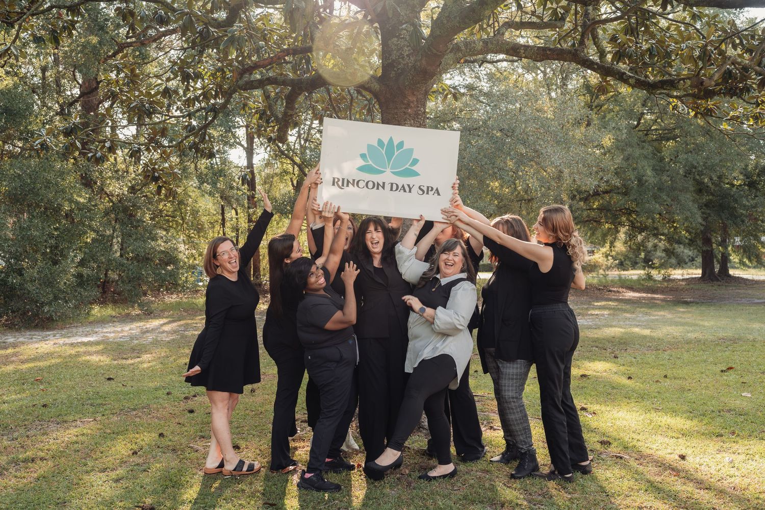 Group of women in black outfits holding a Rincon Day Spa sign together under large oak trees on a sunny green lawn.